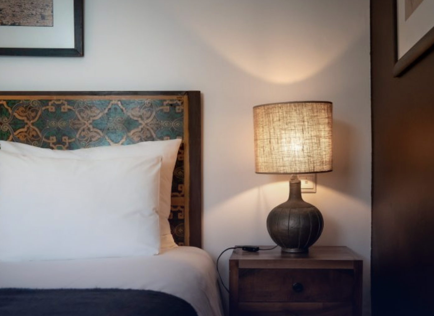 A bedroom scene showing a bed with white pillows and a decorative headboard, a wooden nightstand with a large ceramic lamp, and framed art on the wall.