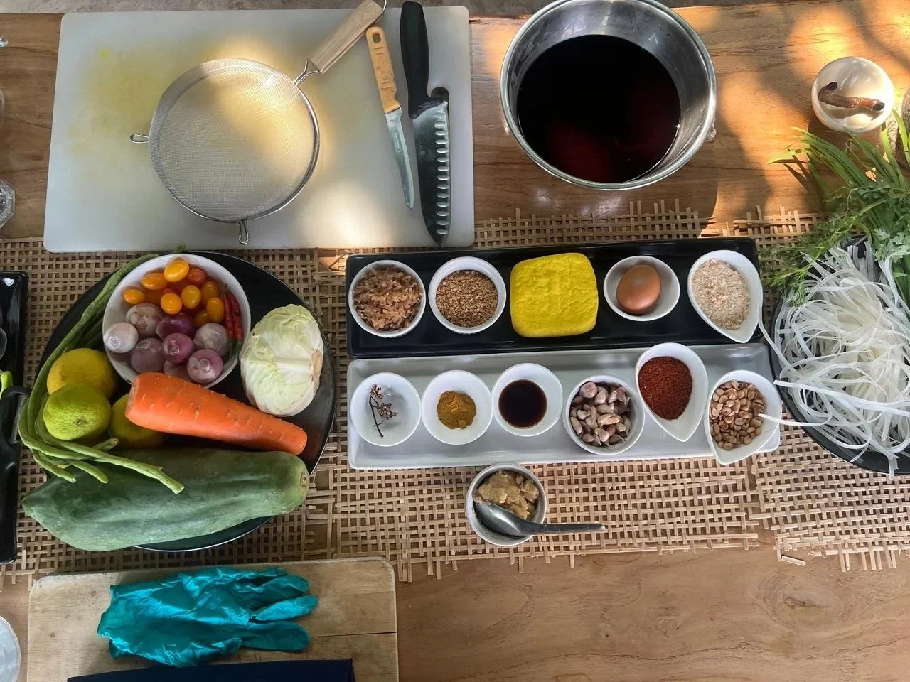 Overhead view of a Thai cooking class station in Chiang Mai showing fresh vegetables, turmeric paste, dried spices, fish sauce, tamarind, rice noodles and herbs laid out in bowls on a woven bamboo mat