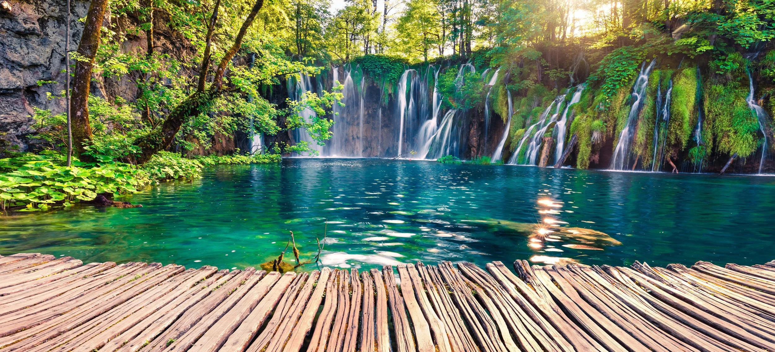 A cascading waterfall flowing into a turquoise lake at Plitvice Lakes National Park, Croatia, with a wooden walkway and lush forest surrounding the emerald water
