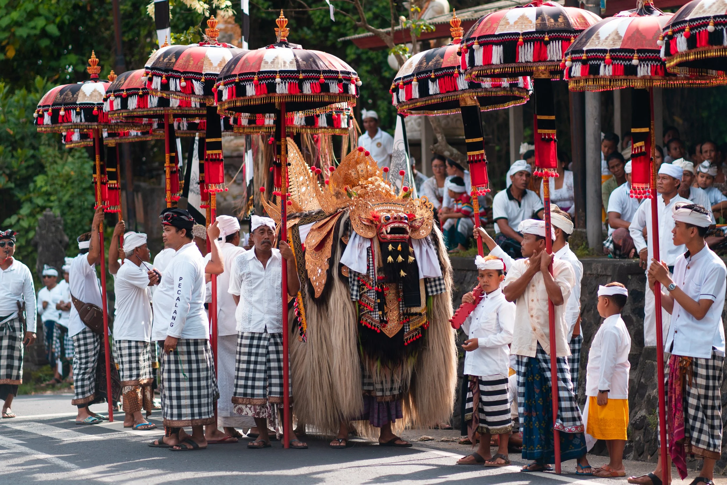 A Barong lion costume carried in a traditional Hindu ceremony procession in Bali, surrounded by men in black-and-white checked sarongs and white shirts holding red and gold ceremonial umbrellas
