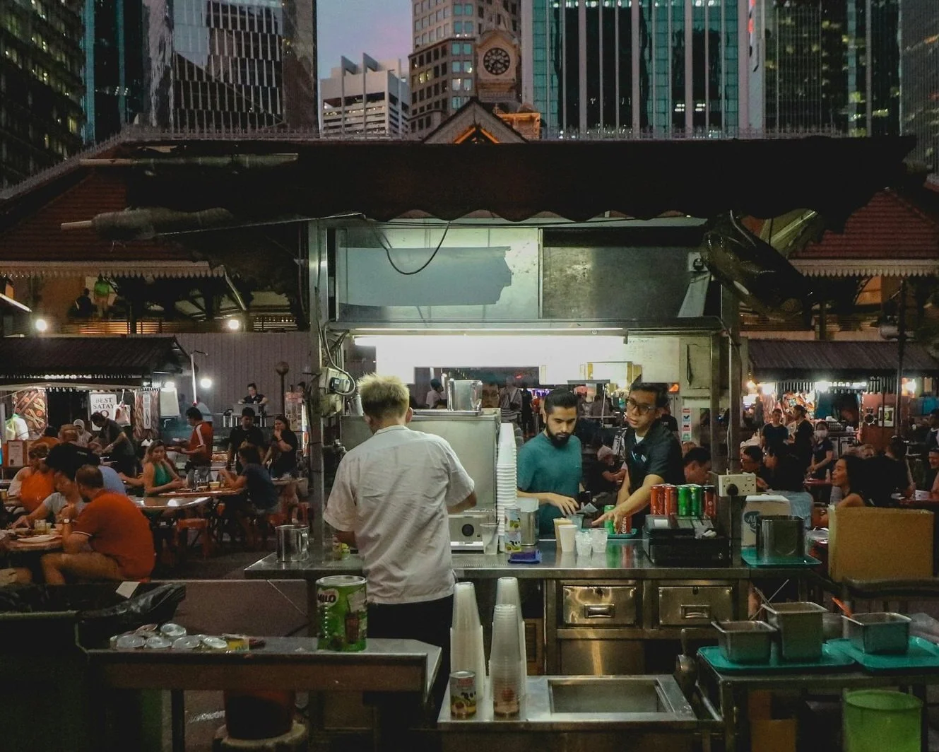A hawker stall chef cooking at a busy open-air hawker centre in Chinatown, Singapore at night, with a crowd of diners, stacked bowls and a lit-up shophouse facade visible in the background