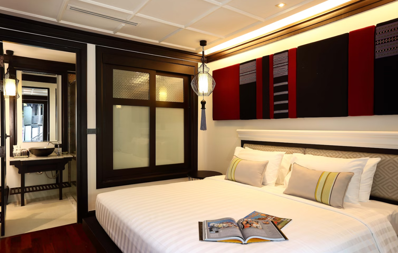 A hotel room with a bed, decorative pillows, colorful wall art above the bed, a bedside table, and a hanging lamp. There is an open magazine on the bed, and a doorway leading to a bathroom with a sink and mirror.