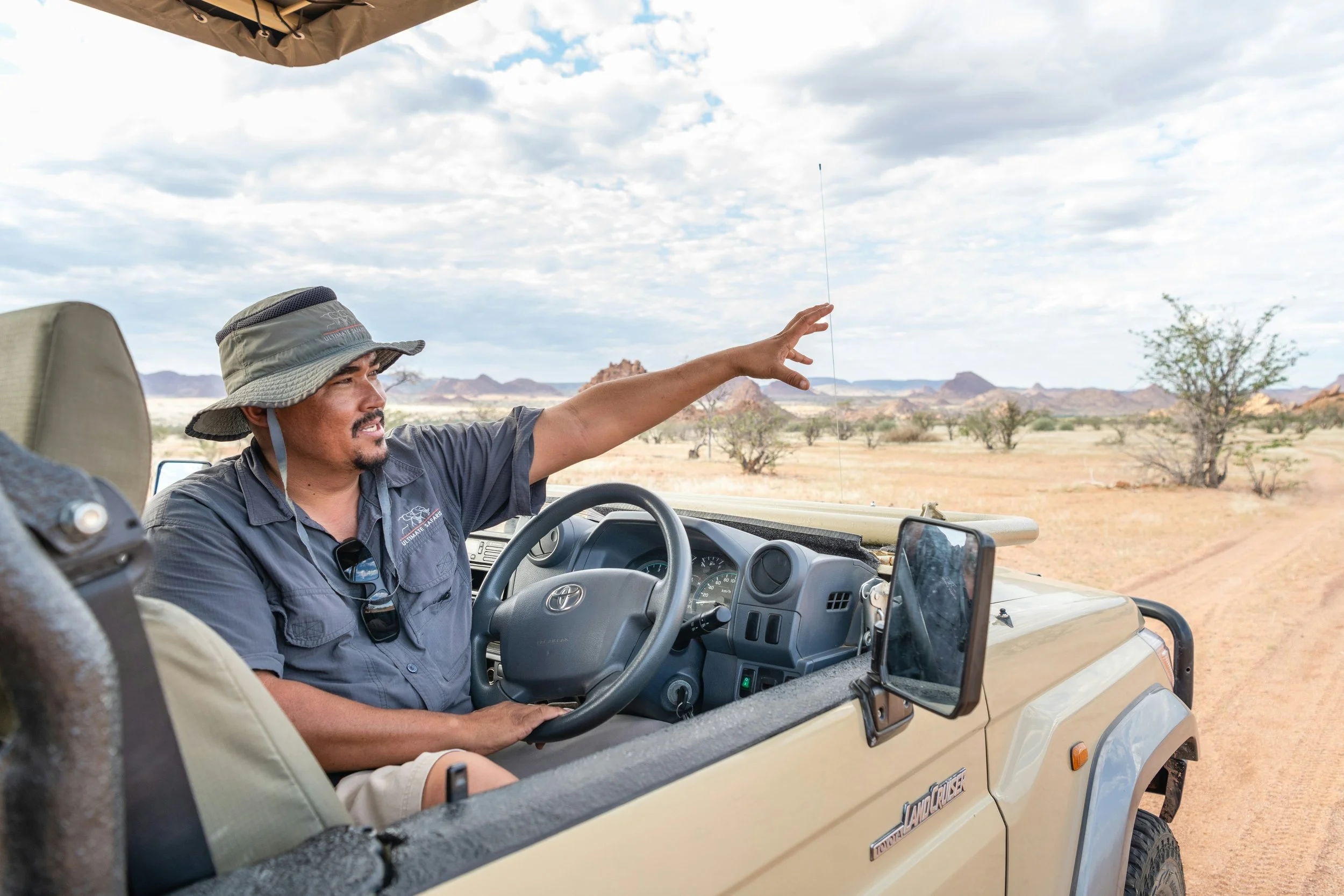 A Namibian safari guide in a bush hat leaning out of an open 4x4 vehicle pointing into the distance across a vast arid Damaraland landscape, with rocky desert hills visible under a partly cloudy sky