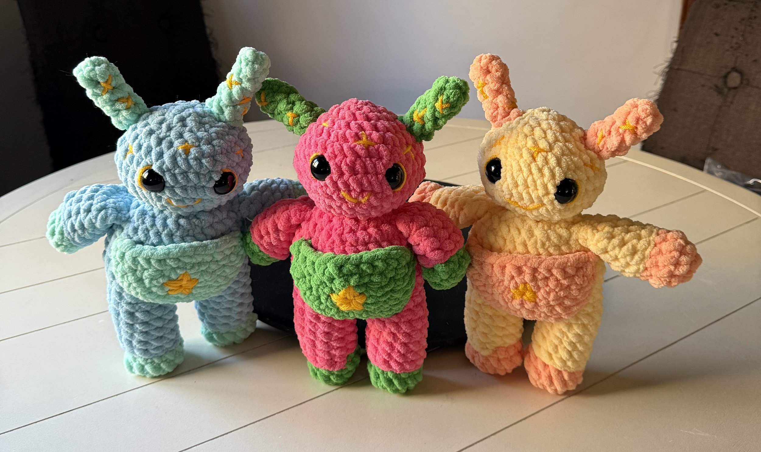 Three plush toys resembling colorful cartoonish creatures with big eyes, antennae, and star decorations, standing on a white table.