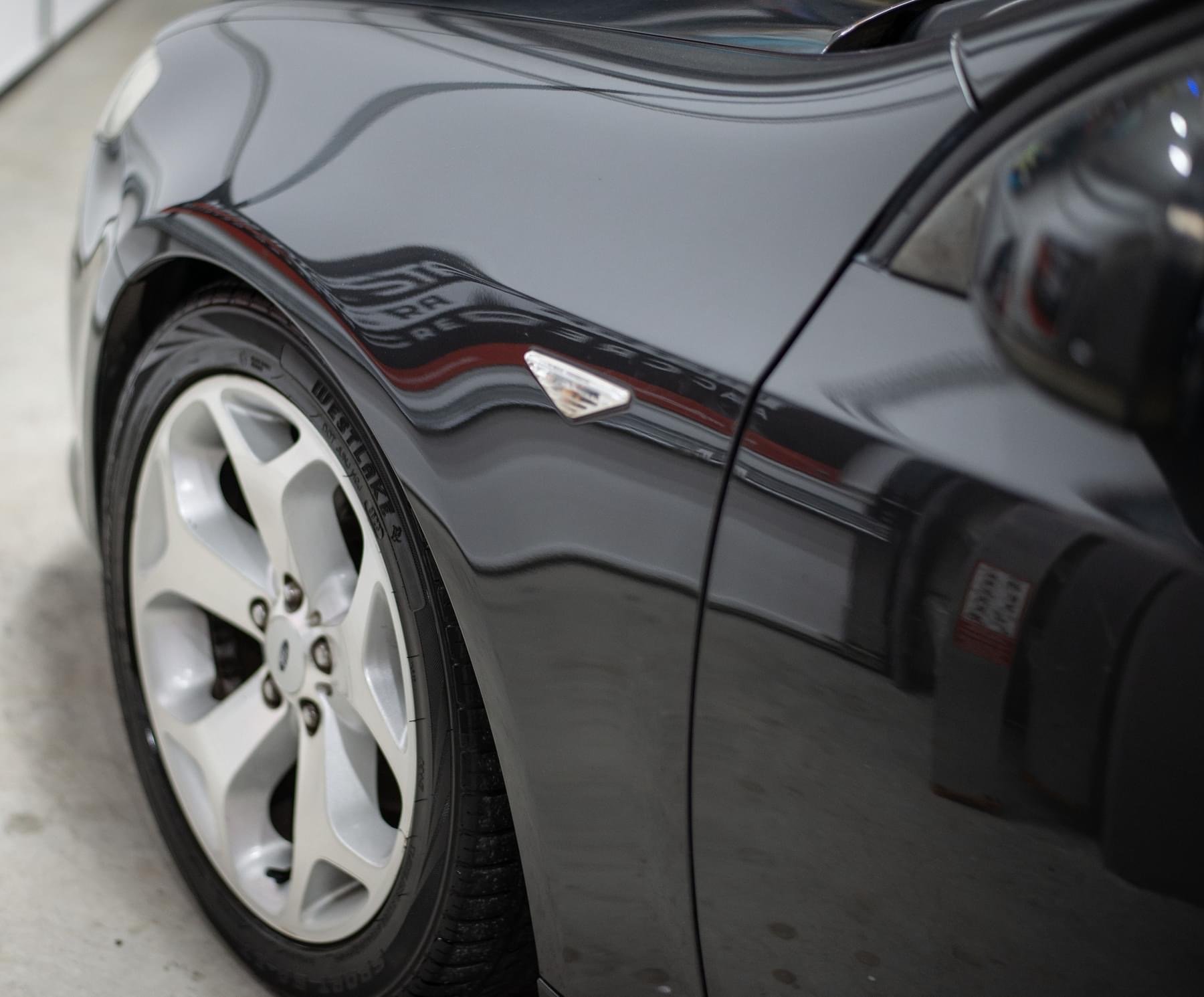 Close-up of a black car's front wheel and fender, showing the vehicle's glossy finish and reflections on the bodywork.