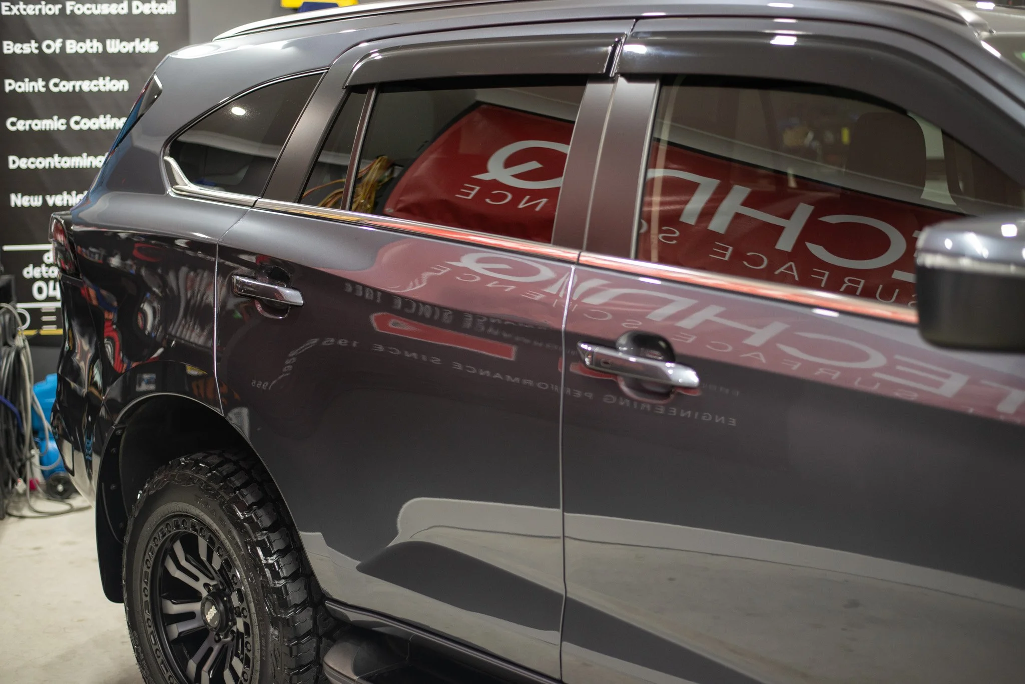 Dark SUV parked indoors with visible reflection and tire detail, surrounded by automotive gear and signage.