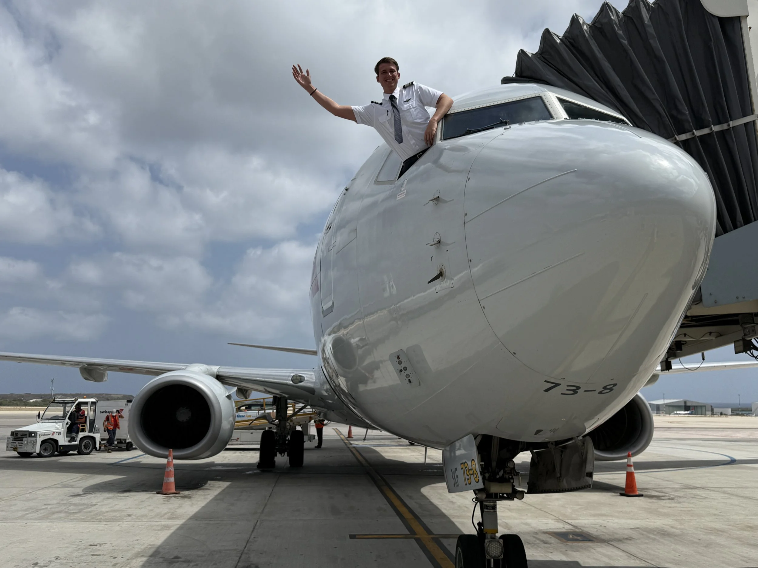 A pilot stands out of the cockpit of a large commercial airplane on the tarmac, smiling and waving. Ground crew and equipment are nearby with traffic cones around the plane, and a cloudy sky overhead.