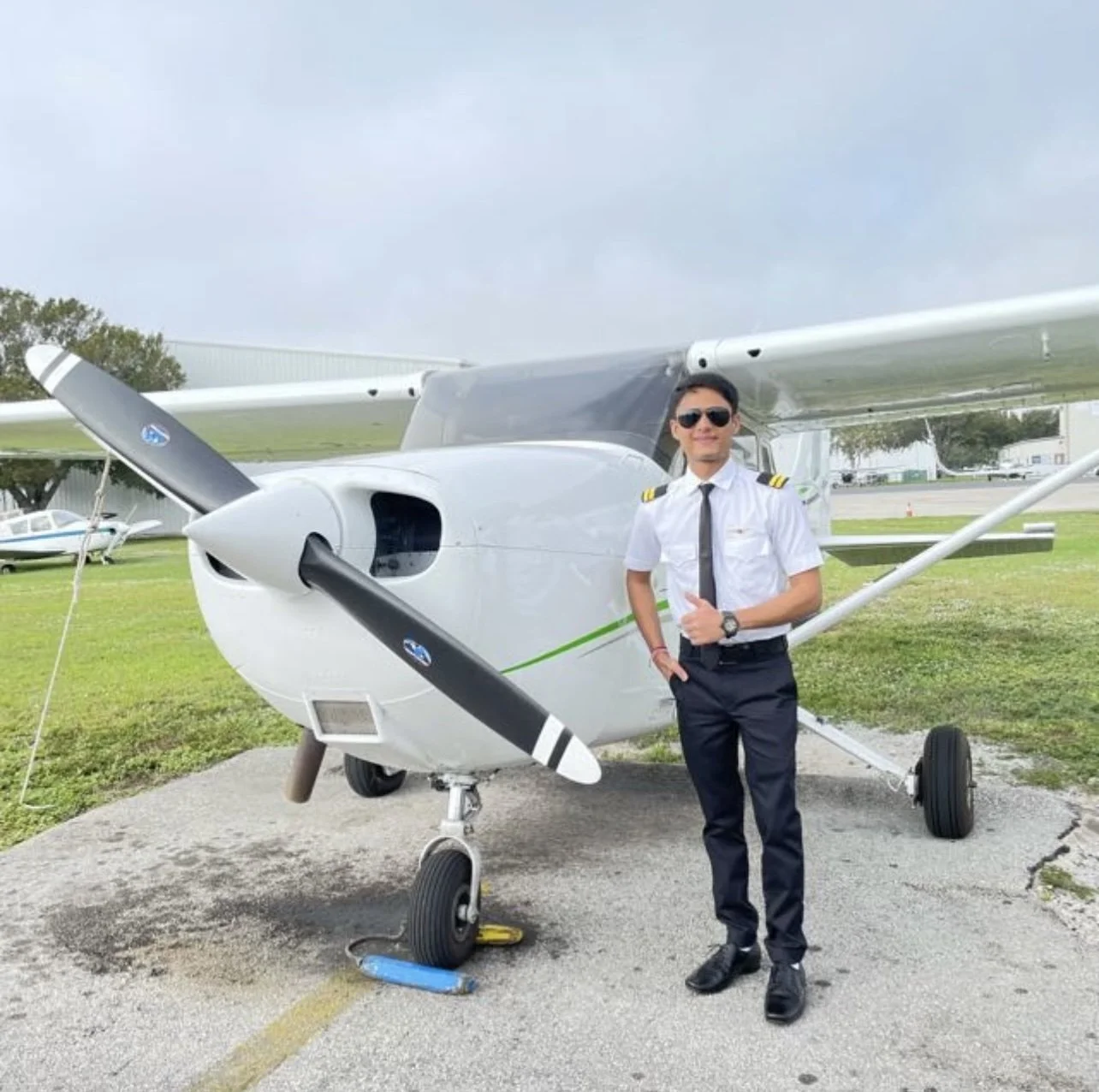Tirth next to a Cessna 172 in a full pilot uniform and sunglasses giving a thumbs up smiling.