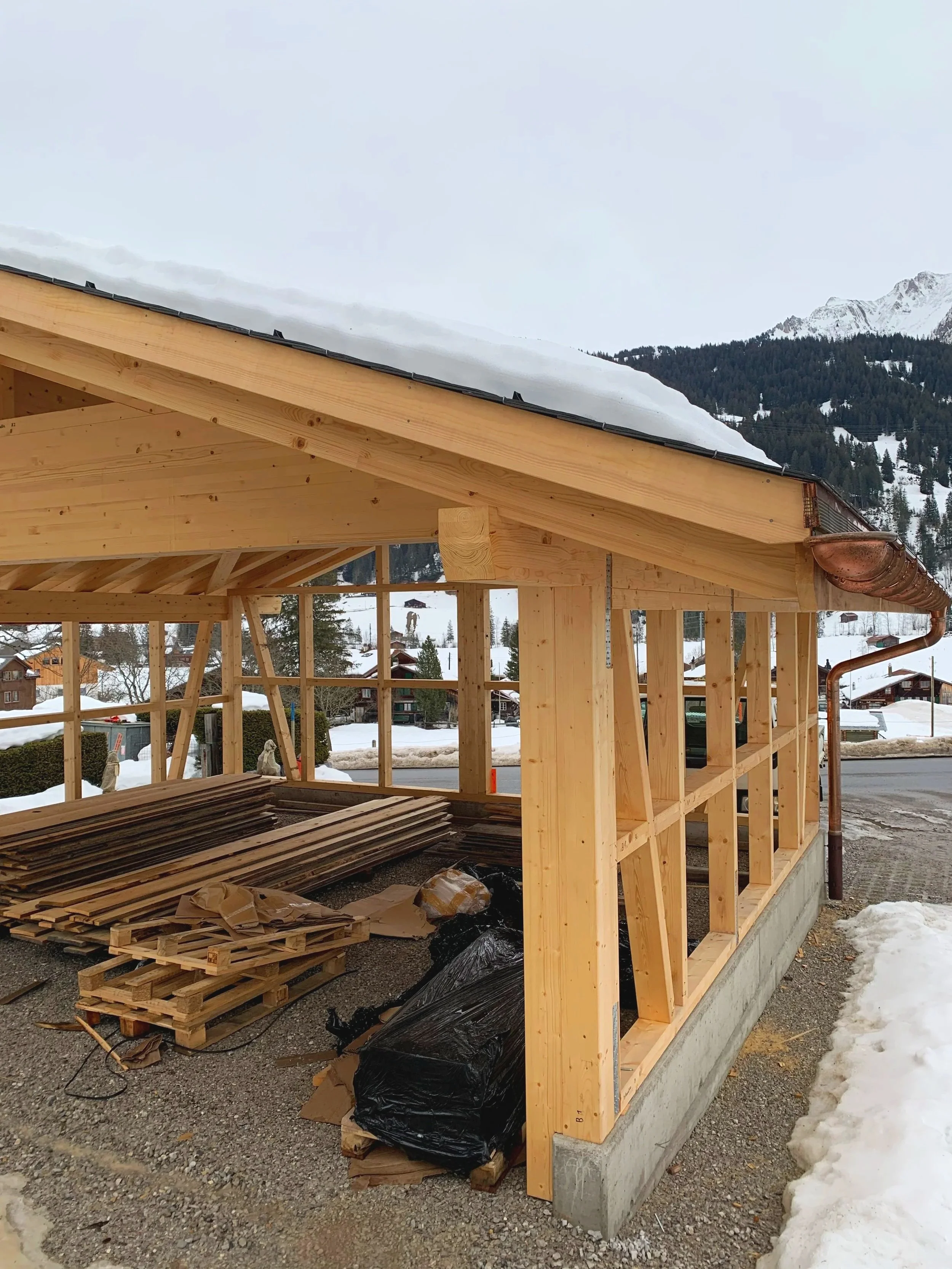 Construction site of a wooden building with a snow-covered mountain in the background. The building's wooden frame is partially completed, with a roof and walls in progress. Building materials and pallets are on the ground.