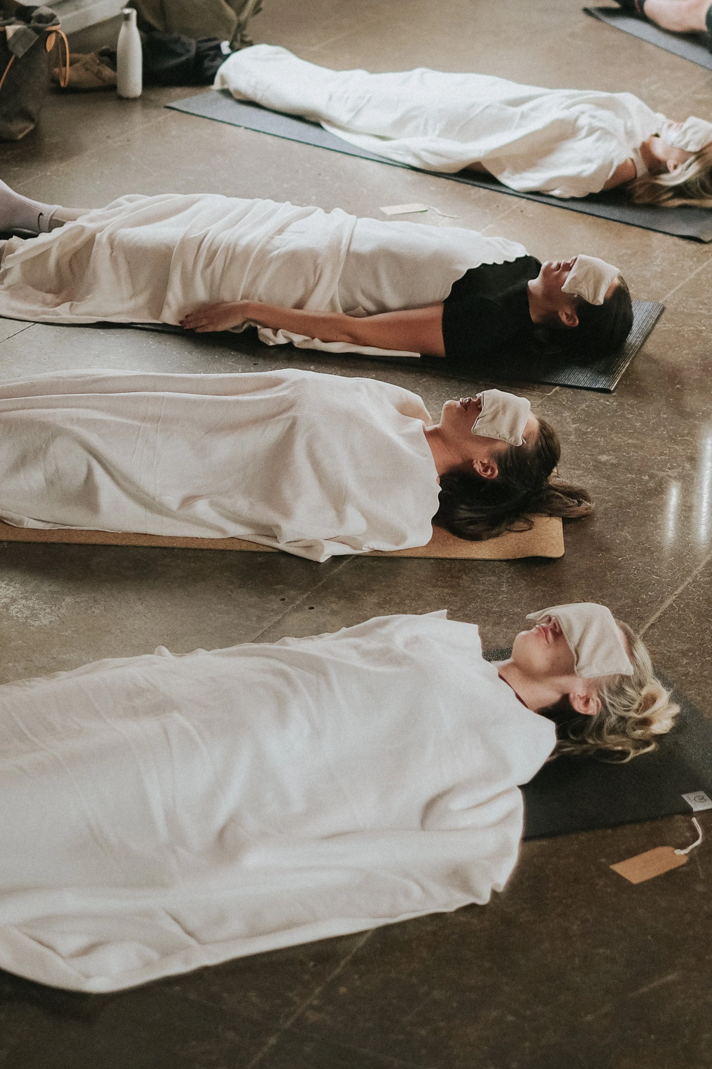 Four women lying on mats on the floor with eye masks during a relaxation or meditation session.