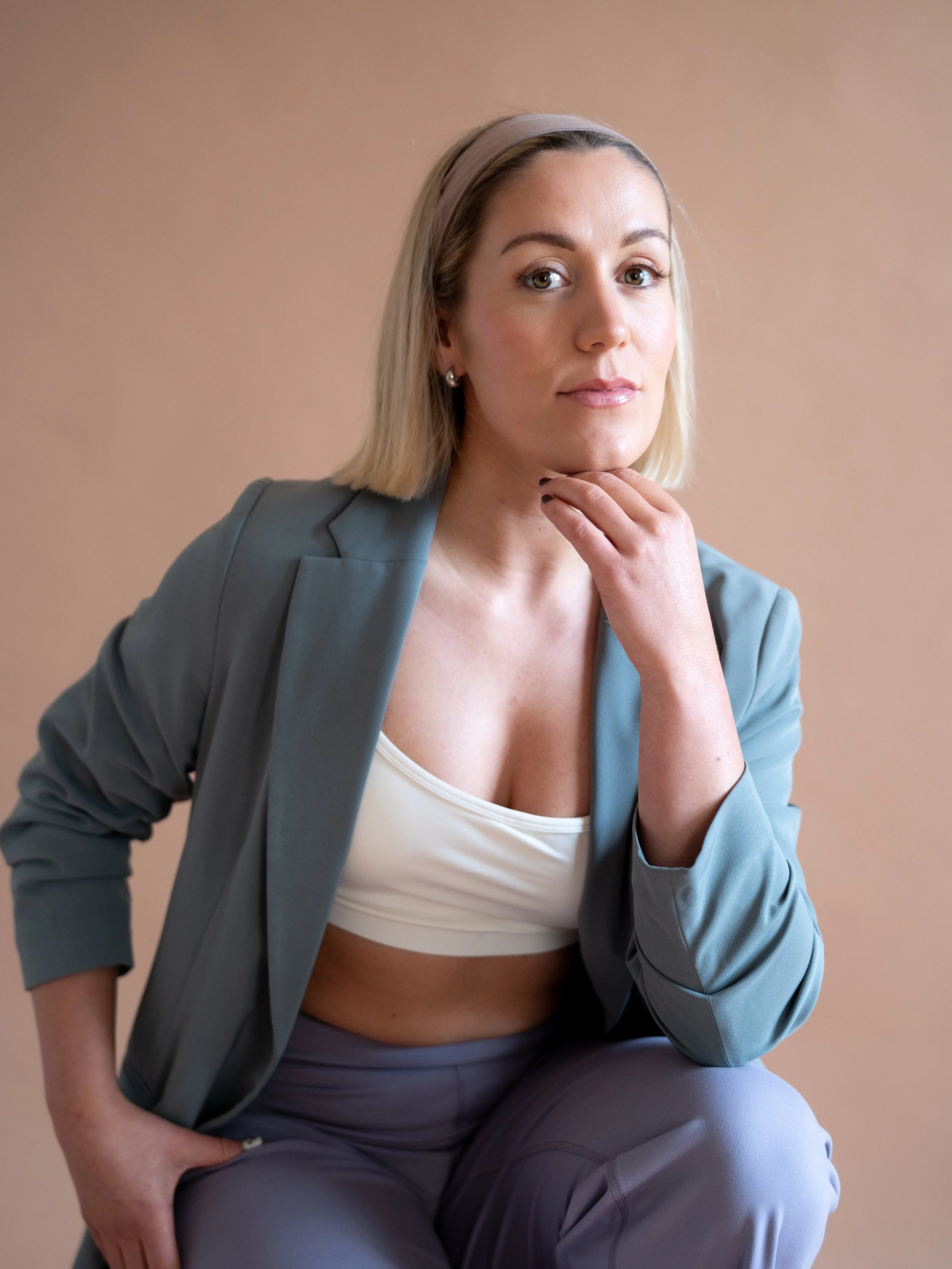 A woman with blonde hair, wearing a light blue blazer and gray pants, sitting against a beige background with her hand resting under her chin.