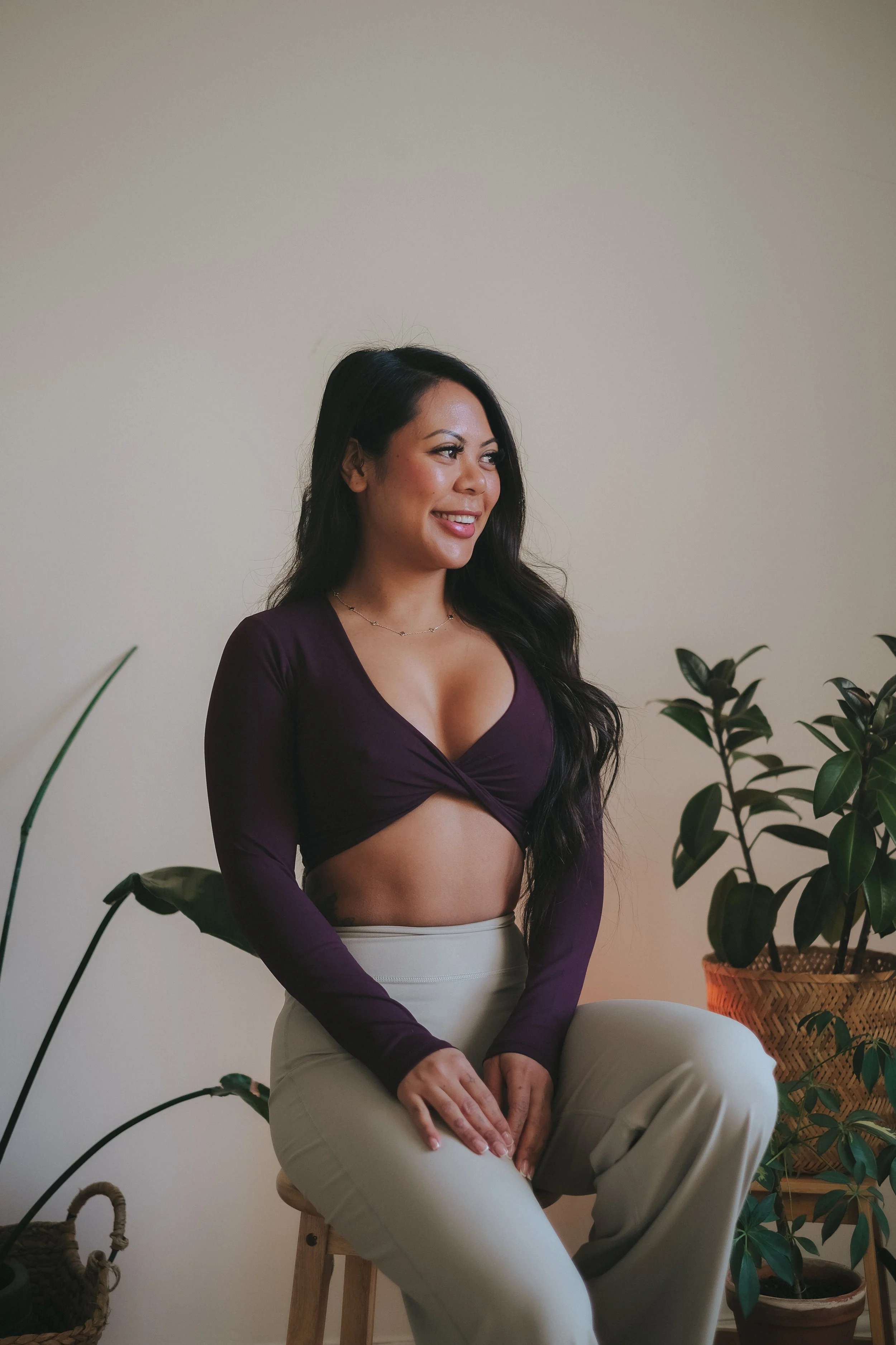 A woman with long dark hair and medium skin tone, smiling and sitting on a stool, wearing a purple long-sleeve crop top and beige high-waisted pants surrounded by green houseplants.