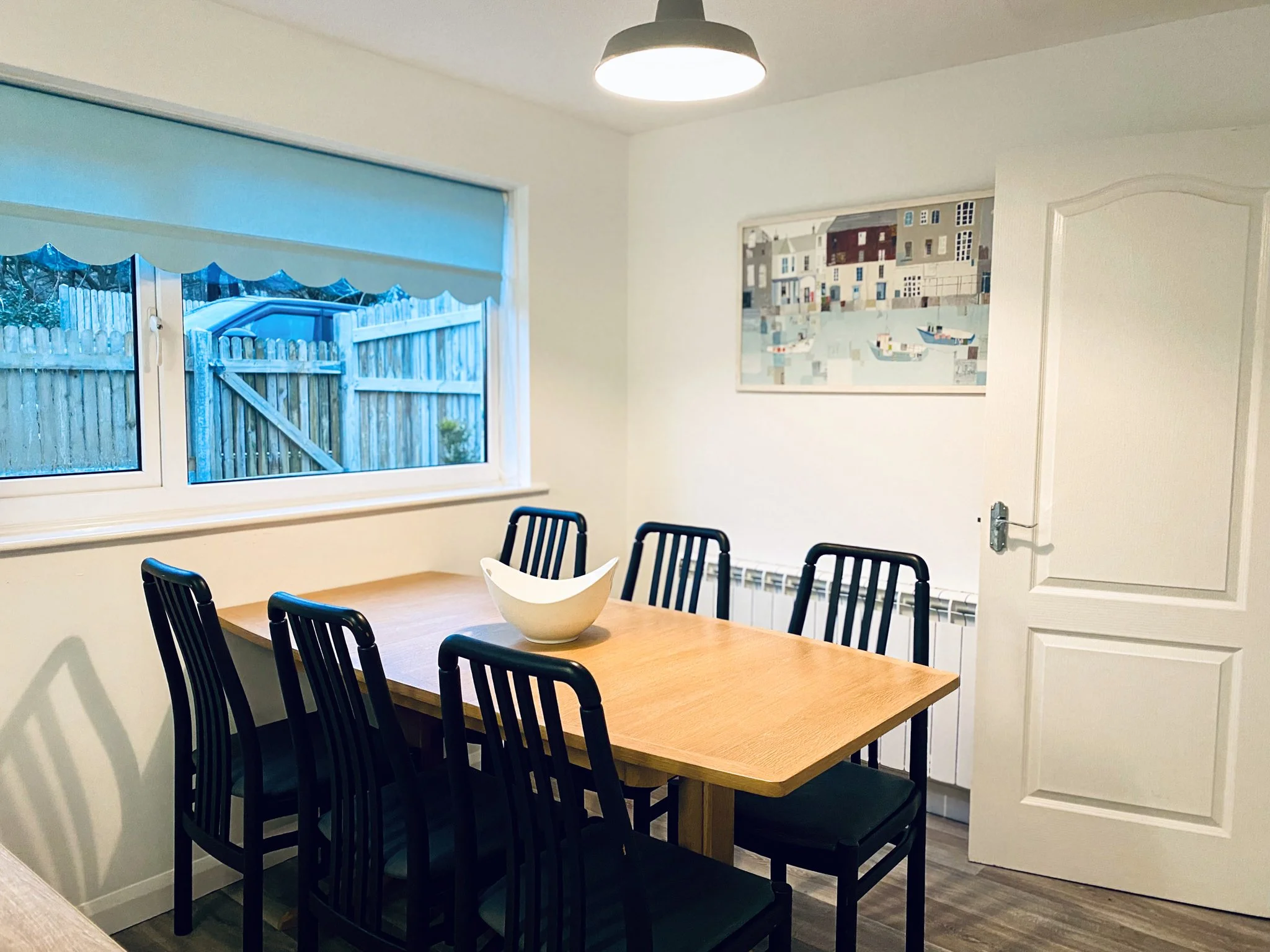 A dining room with a wooden table surrounded by six black chairs, a white bowl in the center, a window with a blue shade overlooking a backyard, a painting on the wall, and a partially open white door.