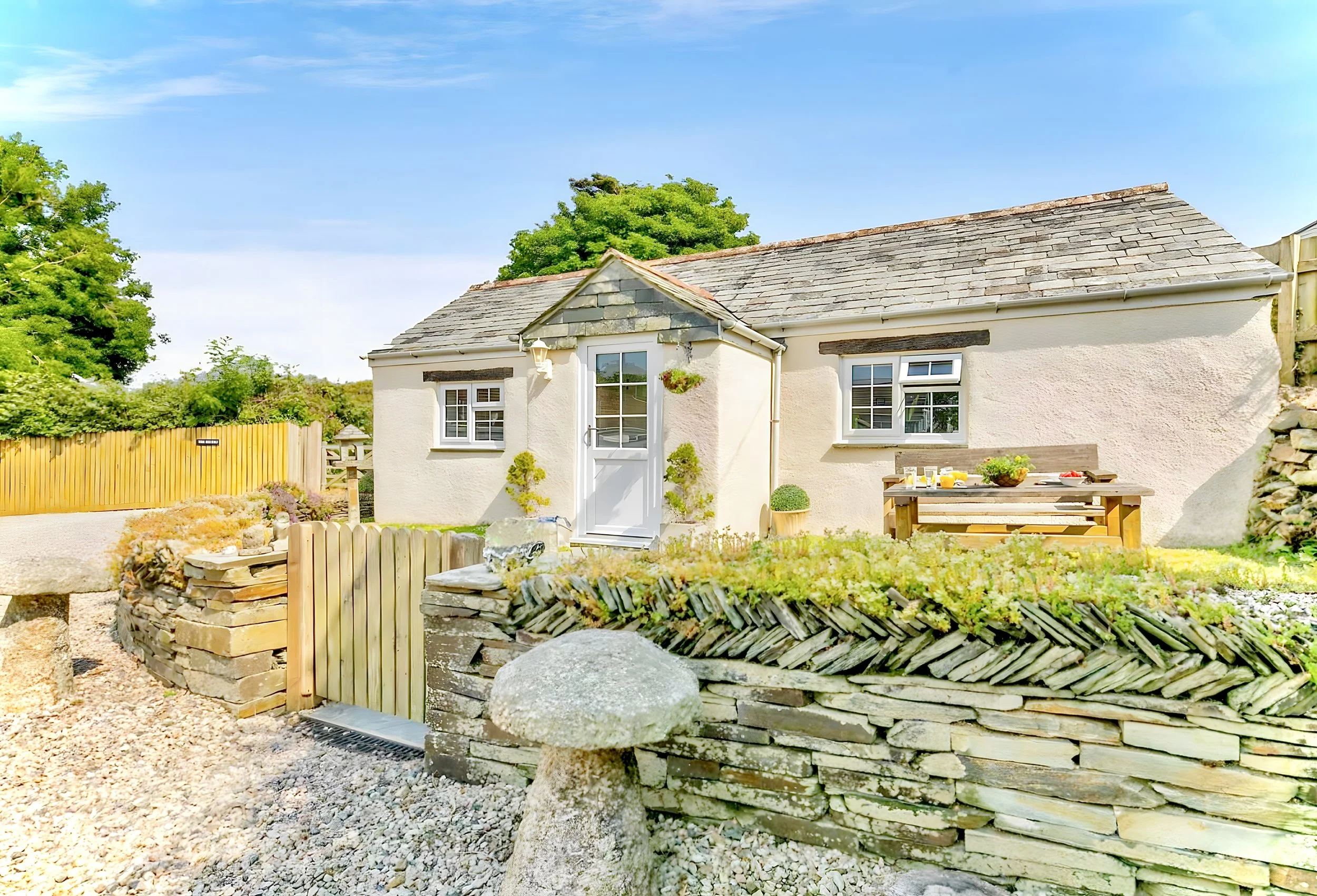 A charming cottage with a stone and stucco exterior, small windows, and a slate roof, surrounded by a yard with a stone retaining wall, wooden fencing, and garden outdoor furniture.