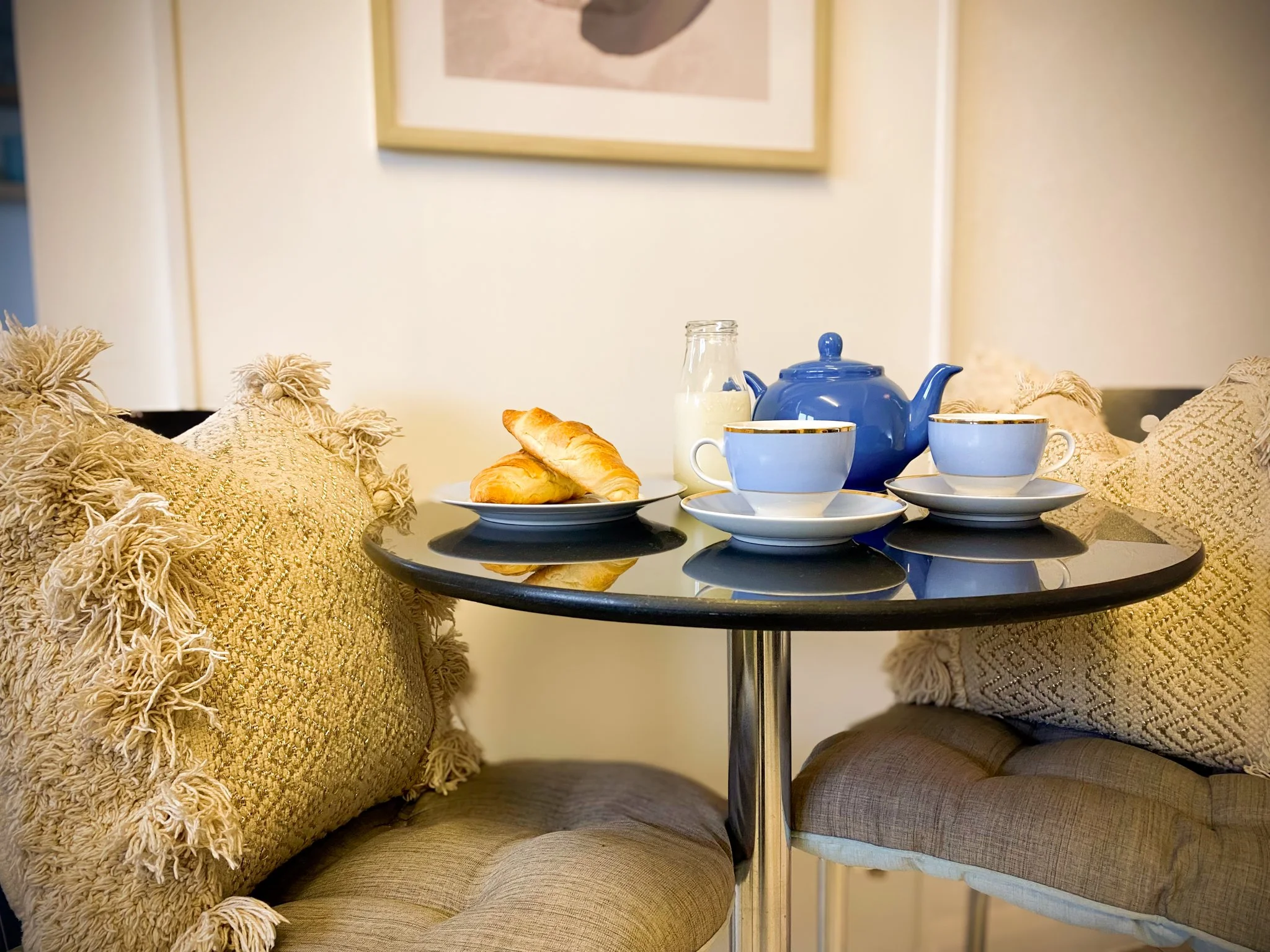 Kitchen table set with a blue teapot, two matching teacups with saucers, a glass of milk, and a plate with two croissants, surrounded by two woven beige pillows on chairs.
