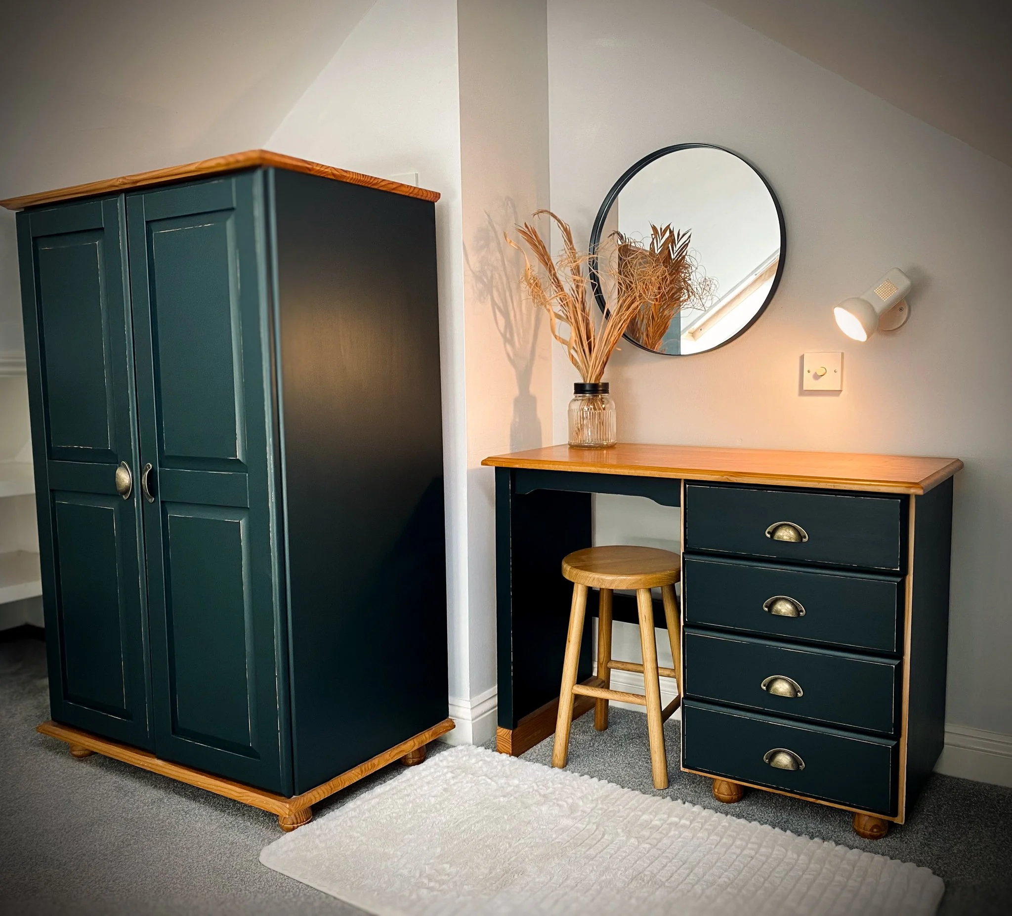 A bedroom corner with a tall dark green wardrobe, a matching dark green desk with four drawers, a round mirror on the wall, a wooden stool, a vase with dried plants, and a white rug on gray carpet.