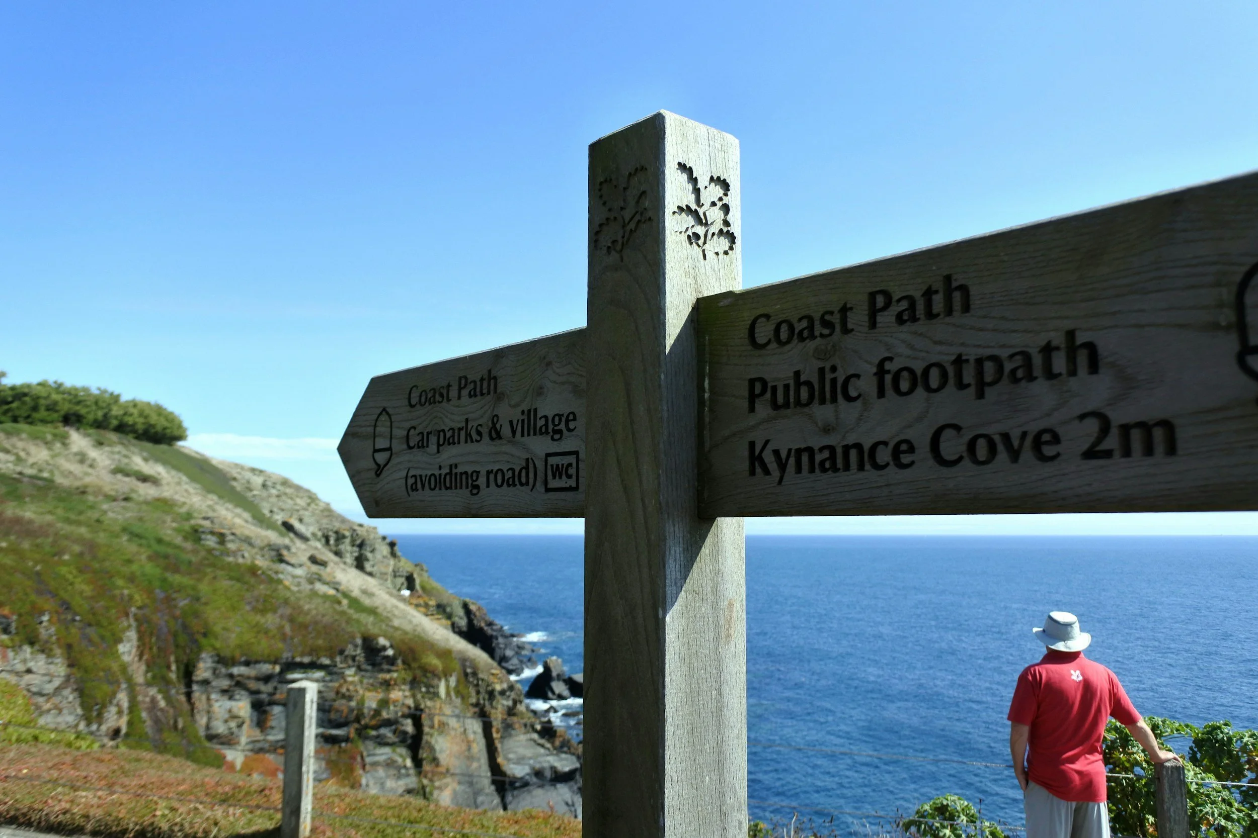 A person wearing a red shirt and white hat standing near a wooden signpost on a scenic coastline, with a cliff, ocean, and clear blue sky in the background.