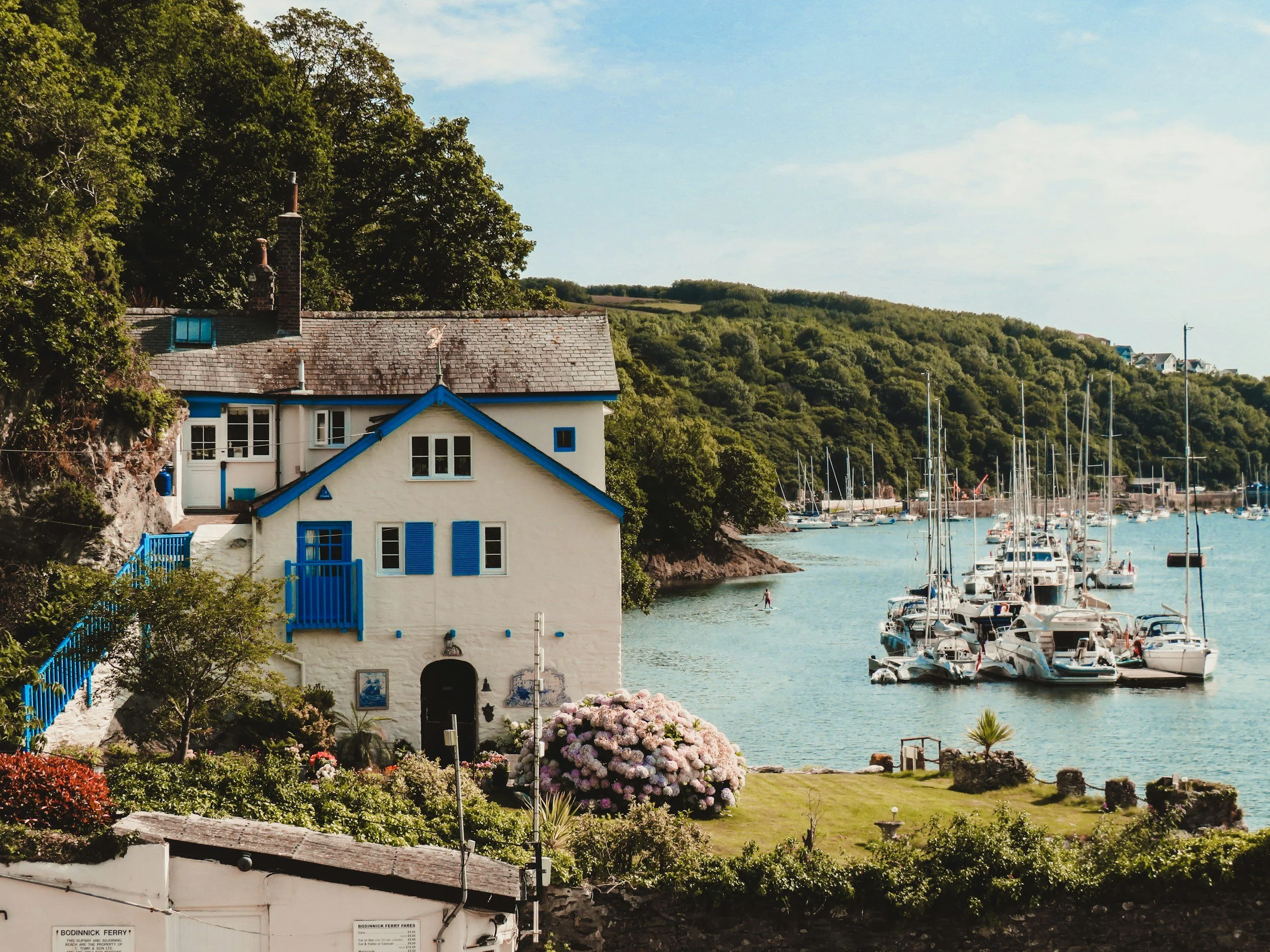 A coastal scene with a white house with blue accents near the water, surrounded by greenery and colorful flowers, with sailboats docked at a marina.