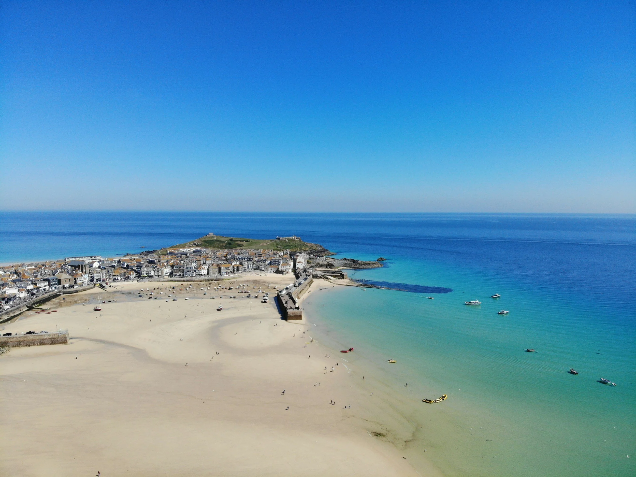 Aerial view of a coastal town with a sandy beach, turquoise water, boats, and a quay, with buildings and houses along the shoreline and a small hill in the distance under a clear blue sky.