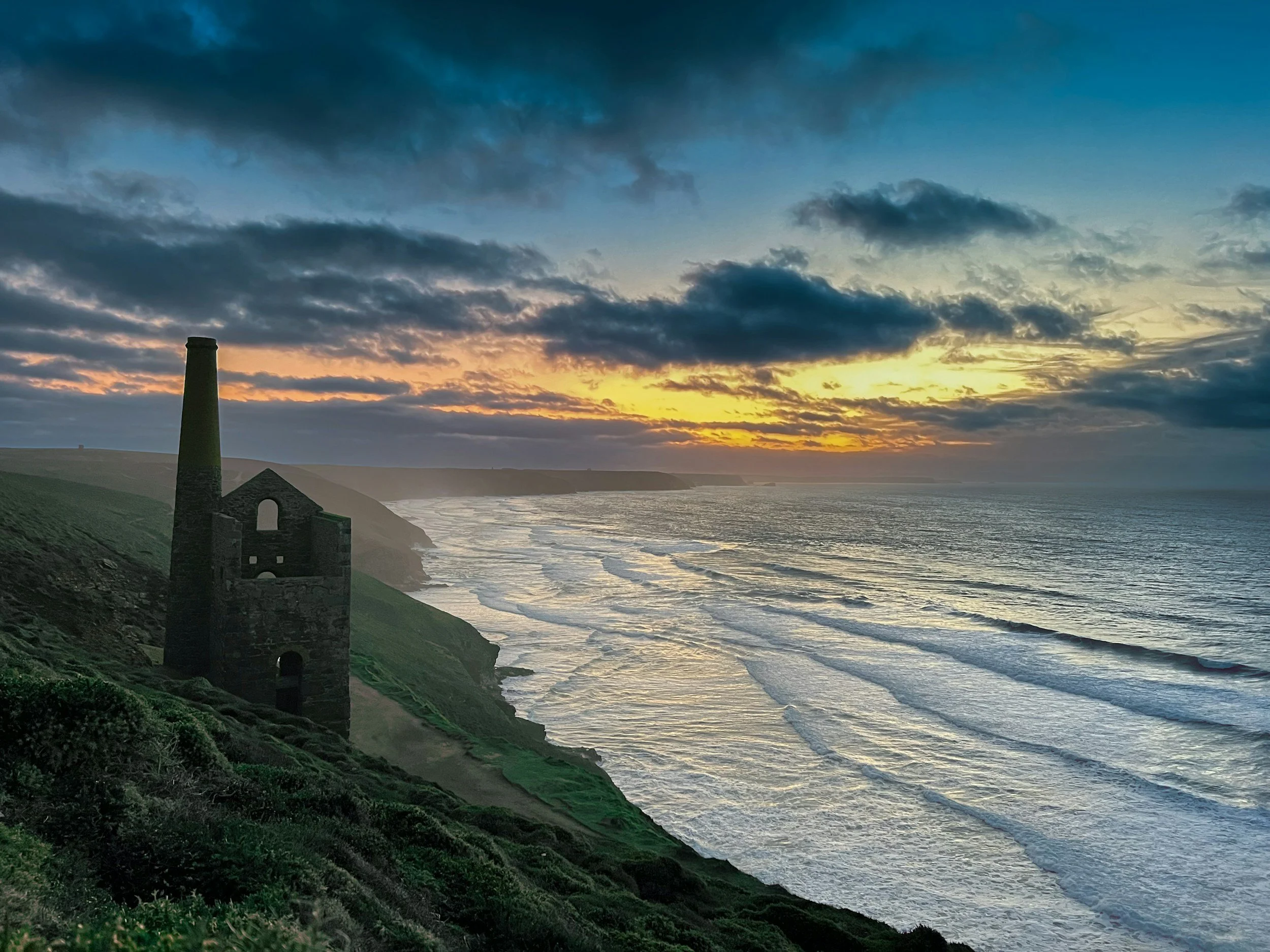 Sunset over the ocean with waves and an old abandoned building on a grassy cliff.