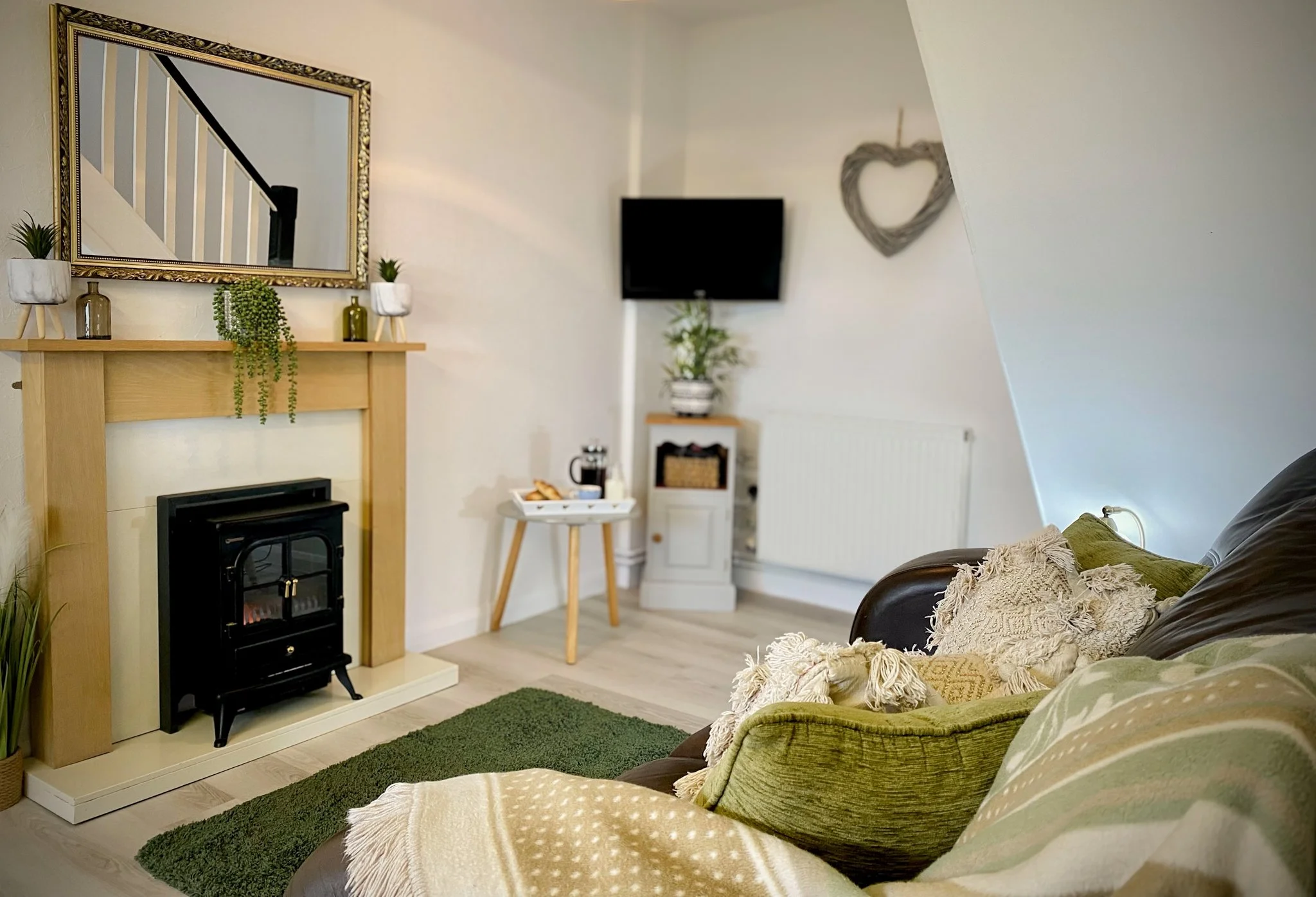 Living room with a green sofa, decorative pillows, a fireplace with a mirror above it, small plants, a television, a small table with snacks and drinks, and wall decorations.