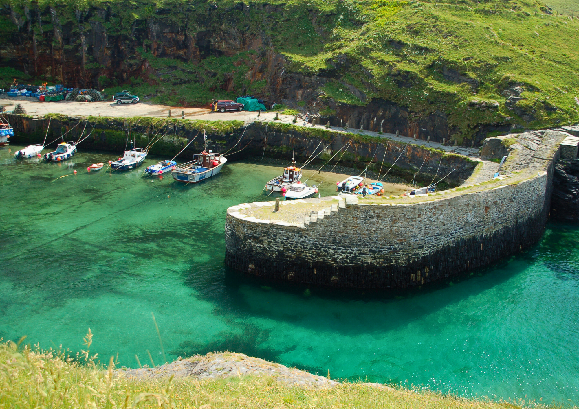 A small harbor with boats anchored along a stone pier, encircled by green hills and rocky cliffs, with a parking area and cars nearby.