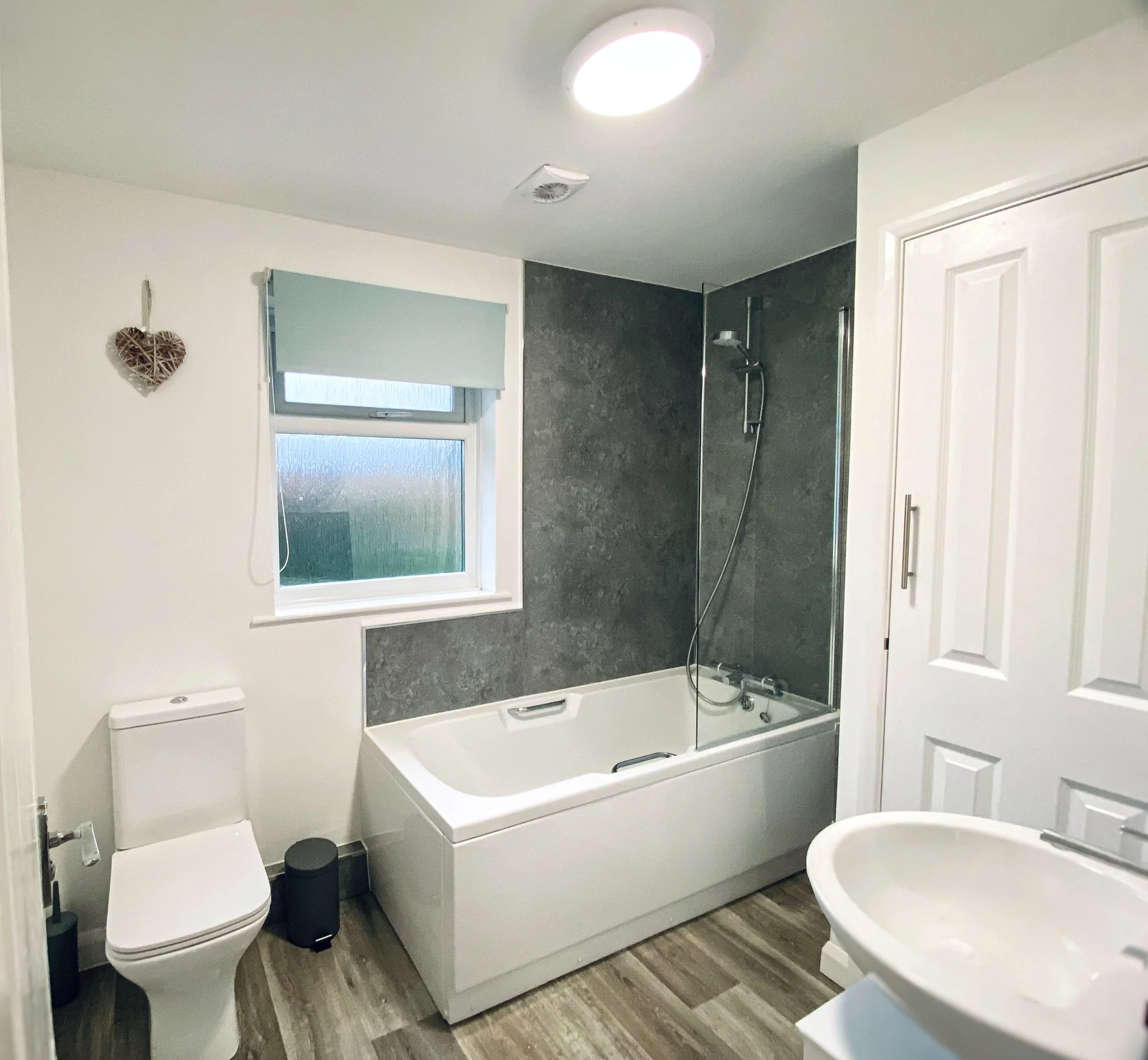 Bathroom with bathtub, shower, toilet, and sink, gray and white color scheme, wooden floor, small window with blinds.