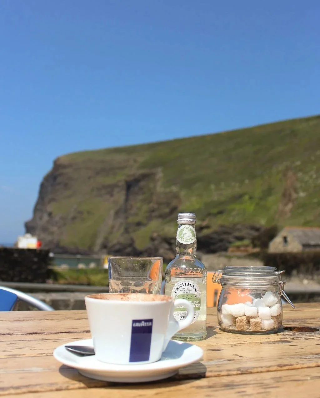 Coffee cup, glass, bottle of gin, and sugar jar on a wooden table with grassy cliffs and blue sky in the background.