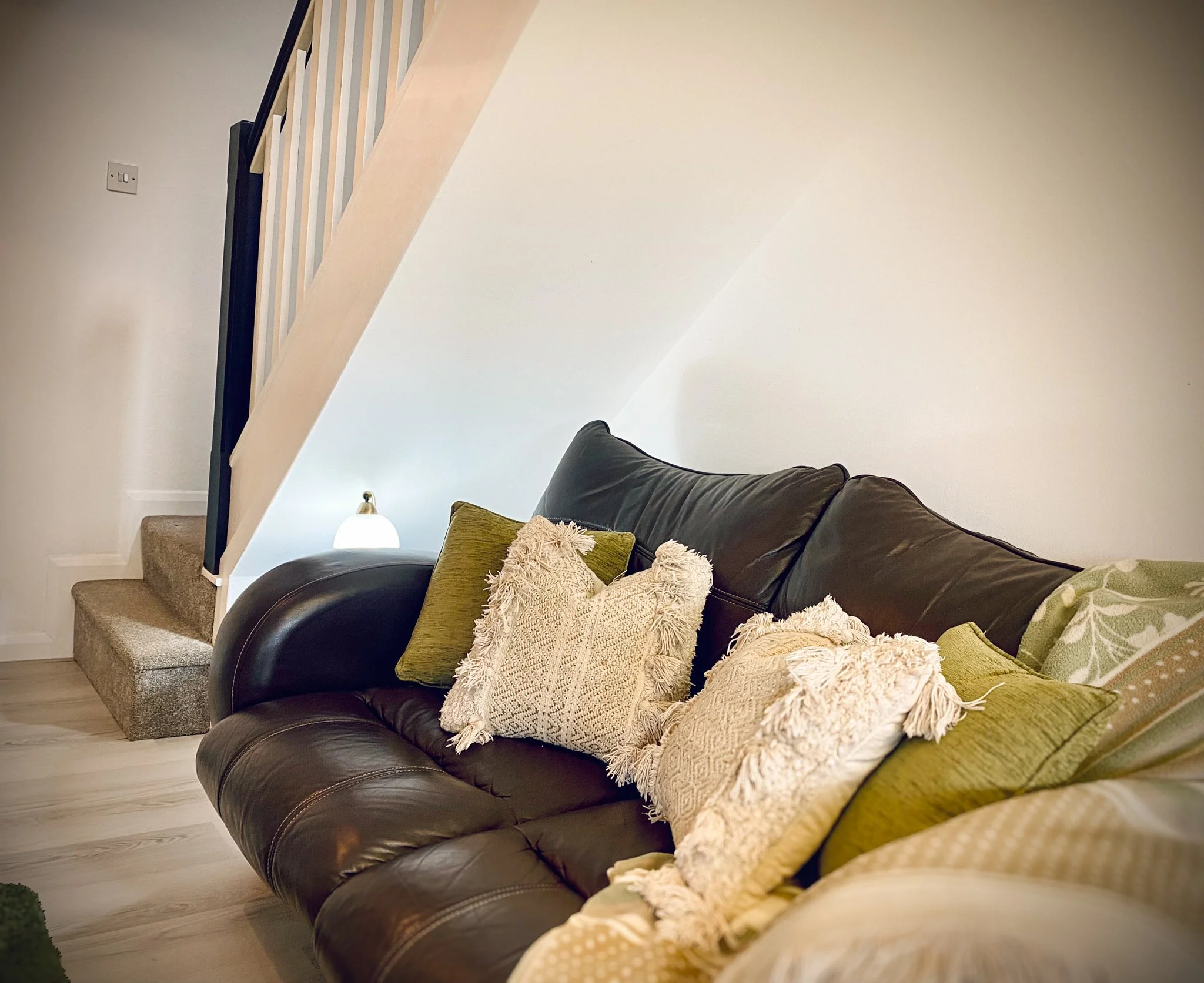 Living room with a dark leather sofa, decorative pillows, a staircase with carpeted steps, and a small lamp on a shelf.