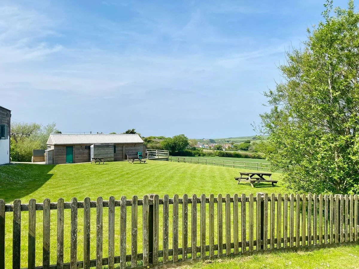 A grassy field with several picnic tables, a small wooden building, trees, a wooden fence in the foreground, and a distant view of rolling hills under a partly cloudy sky.