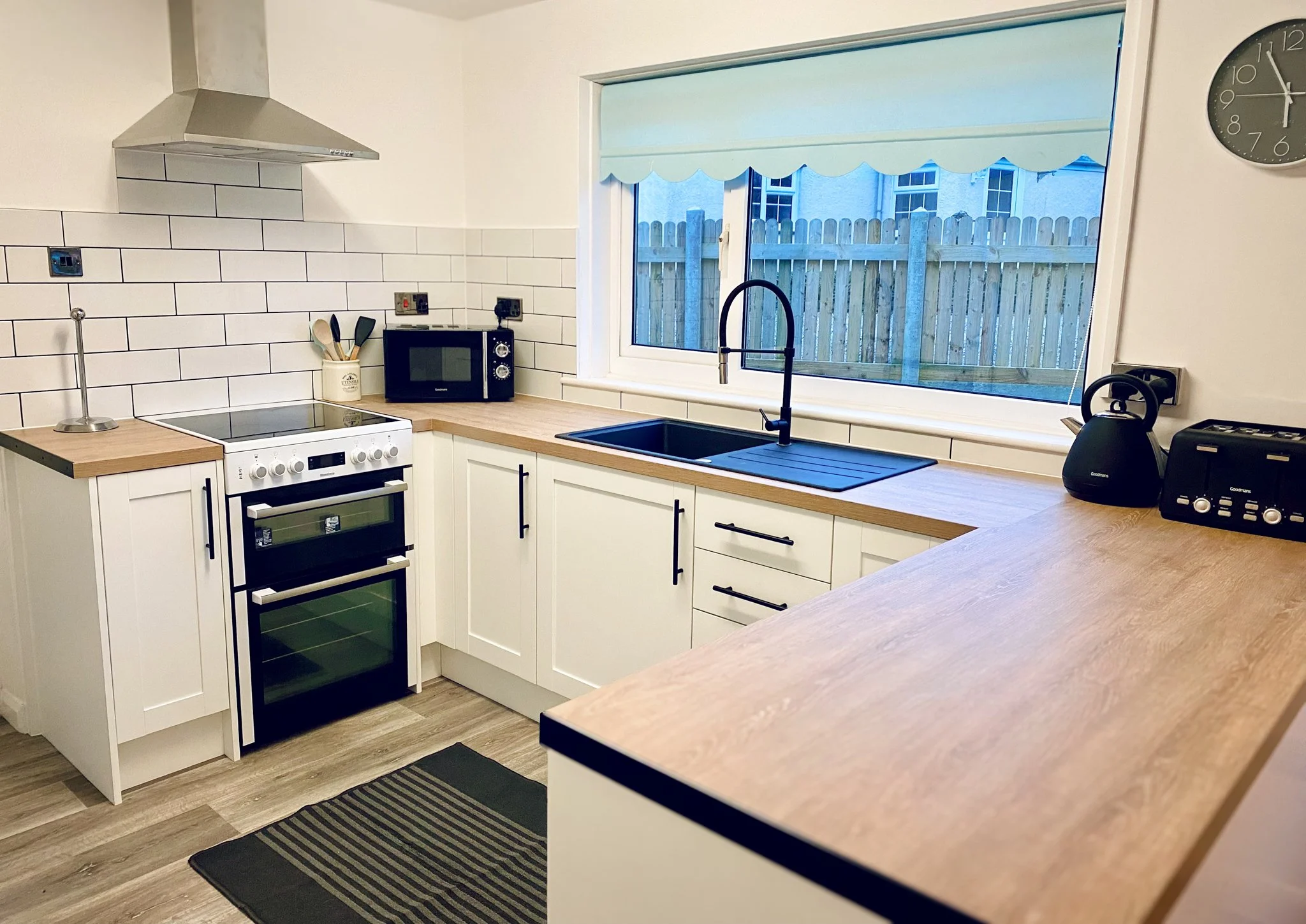 Modern kitchen with white cabinets, wooden countertops, black handles, white subway tile backsplash, stainless steel range hood, black electric stove, microwave, kettle, toaster, black clock, and large window overlooking a fenced backyard.