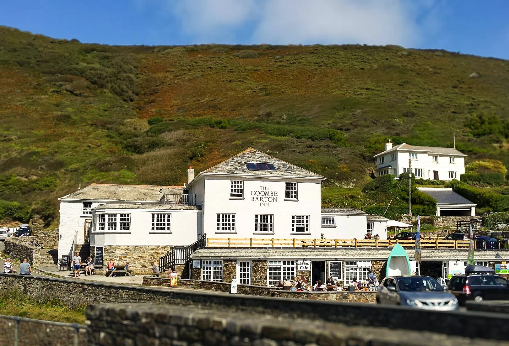 A white multi-story building called The Coombe Barton Inn situated near a hillside with green vegetation. There are people sitting outside on benches and cars parked in front.