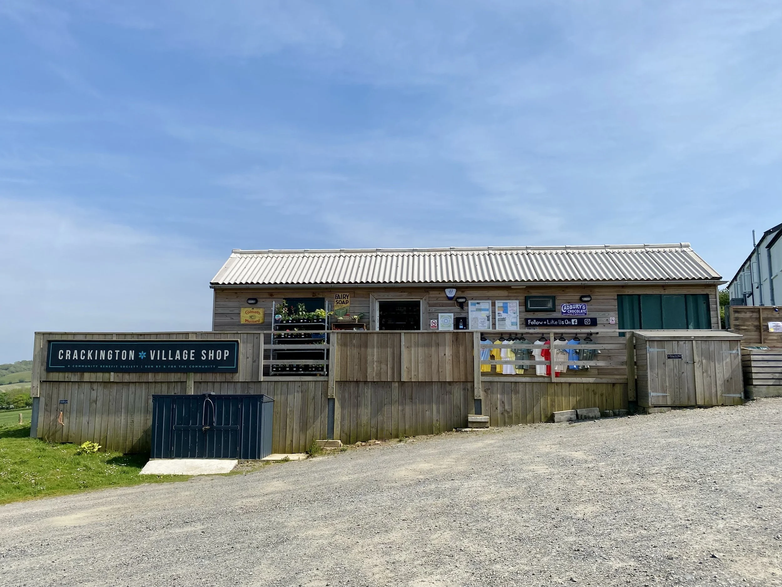 The Crackington Village Shop, a small wooden building with a metal roof, situated on a gravel road with green grass and a rural landscape in the background, under a blue sky.
