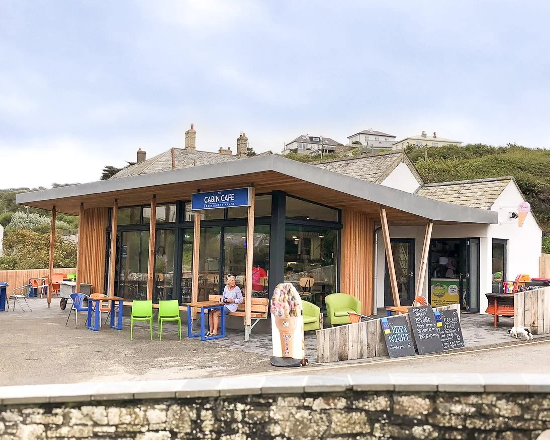 Small modern cafe with outdoor seating, colorful chairs, and a sign that reads 'Cabin Cafe Crackington Haven.'