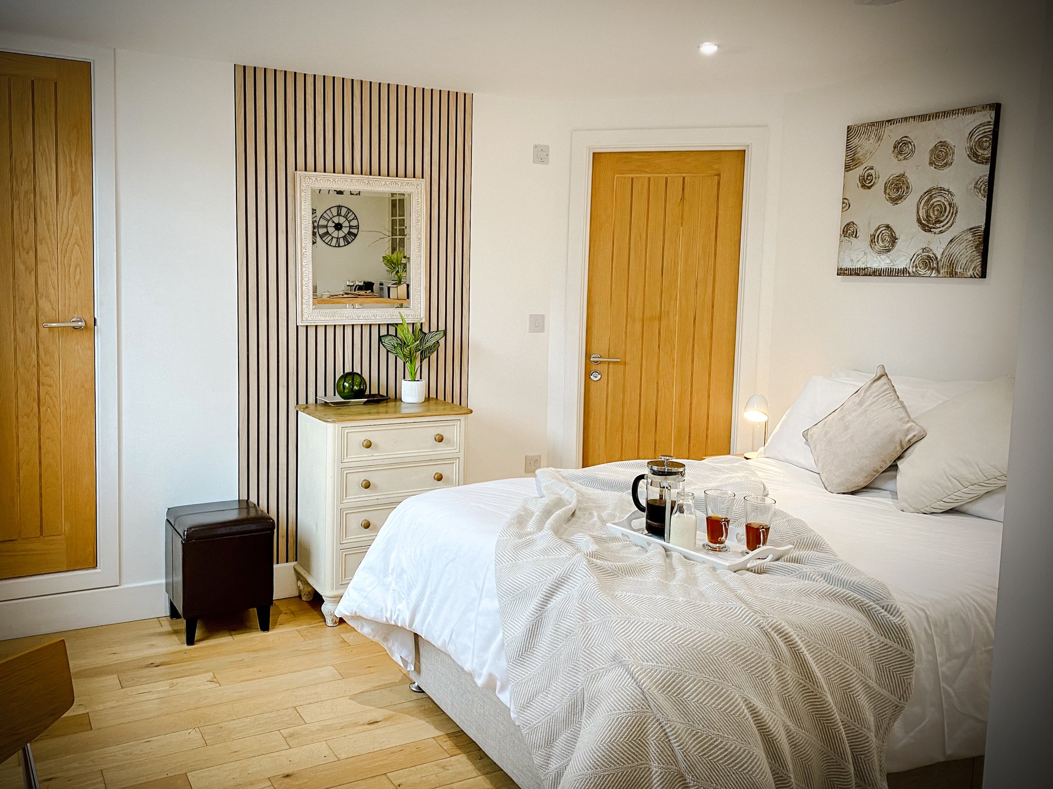 A cozy bedroom with a white bed, a tray with a coffee pot, glasses, and cups, a bedside lamp, a white dresser with a plant and decorative object, a large mirror, wooden doors, and abstract wall art.