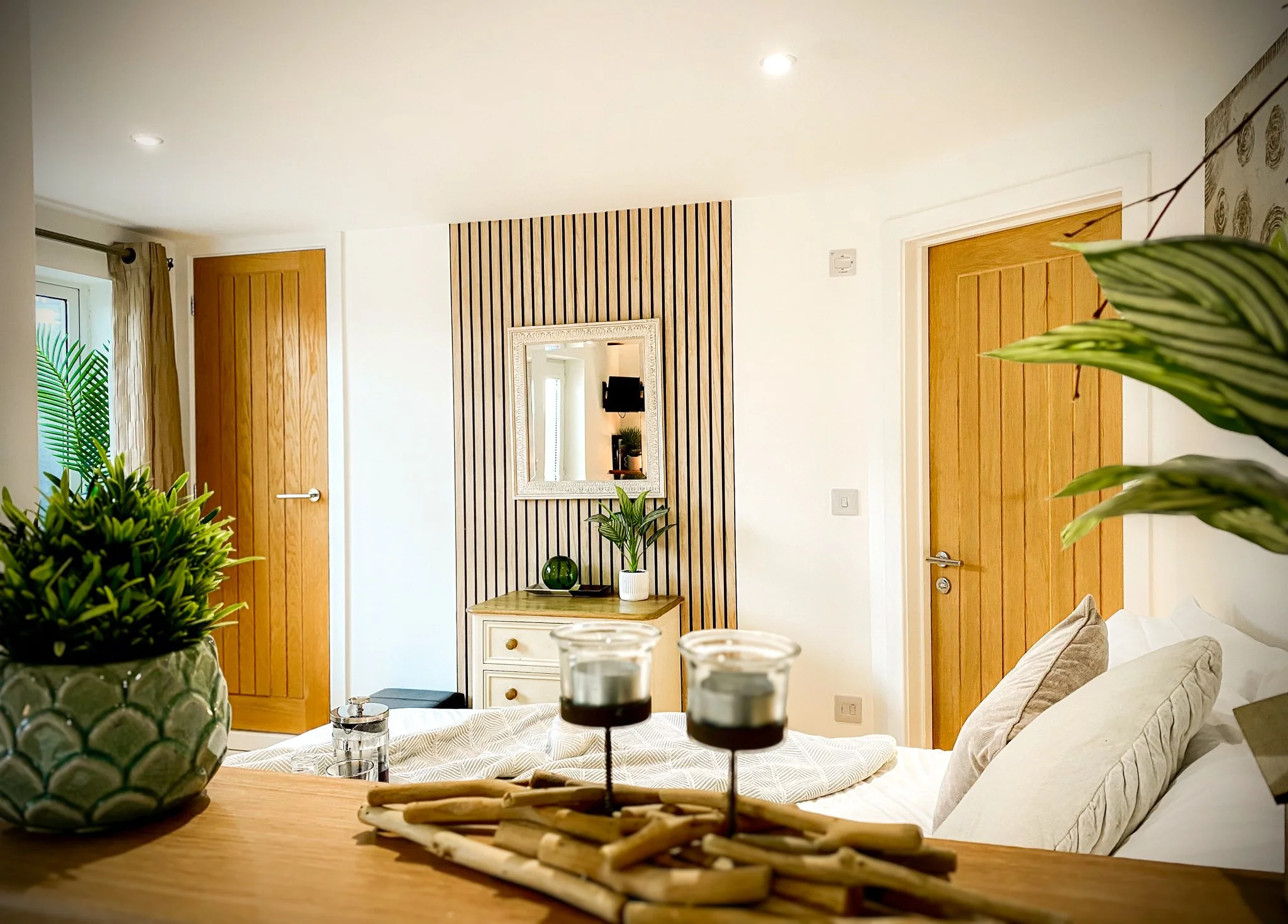 Interior of a modern bedroom with wooden doors, a mirror on a wooden slat wall, a small drawer with a potted plant, candles, and decorative items on a table, pillows on the bed, and plants in the foreground.