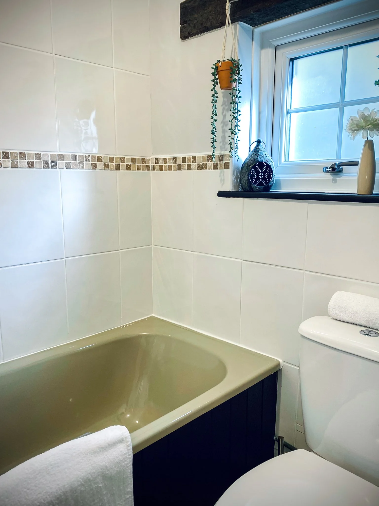 Bathroom with white tiled walls, a beige bathtub, a toilet, a window with a black windowsill, and decorative items including a hanging plant, a vase with a white flower, and a lantern.