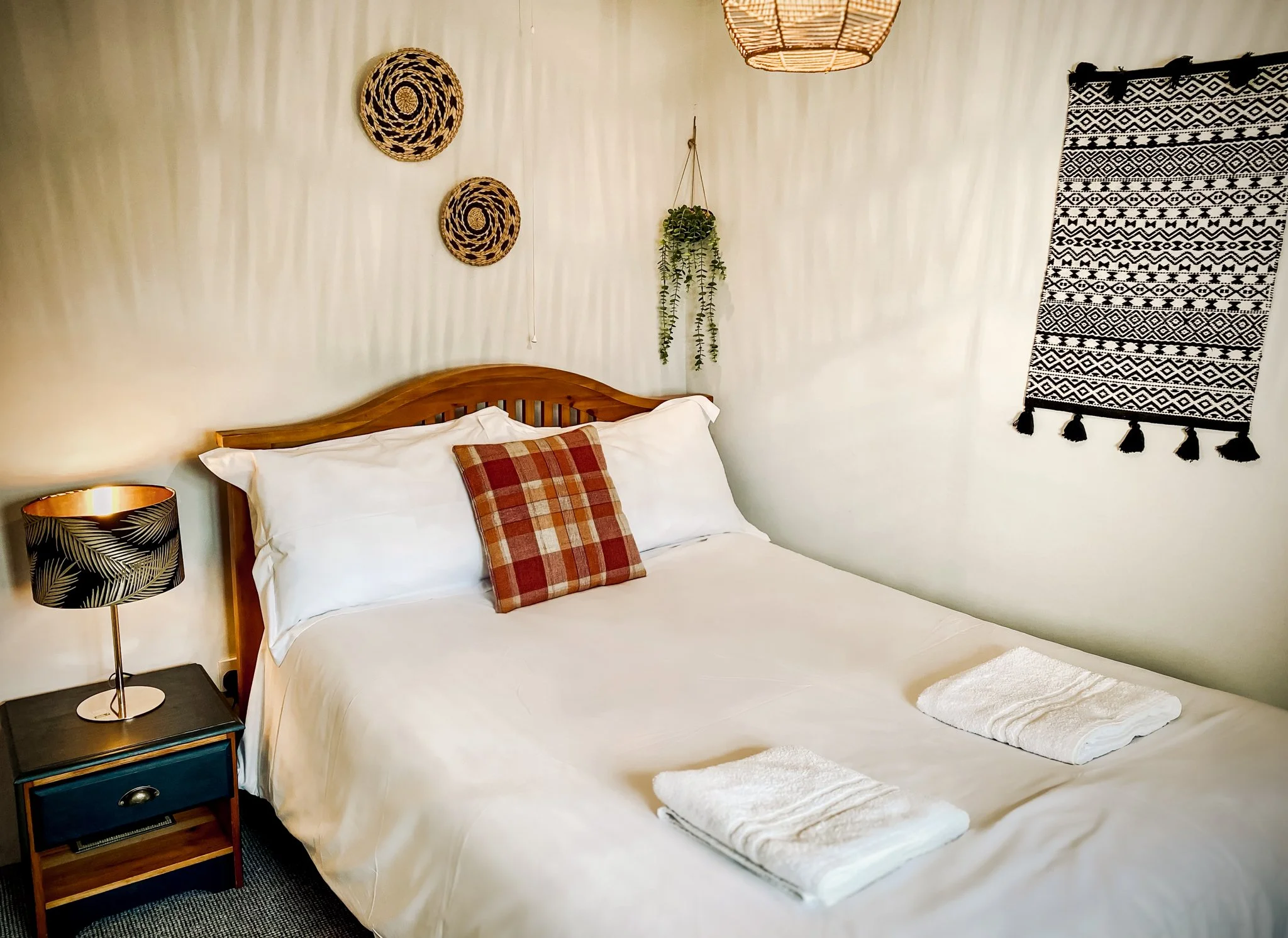 A cozy bedroom with a wooden bed frame, white bedding, a plaid throw pillow, and two neatly folded white towels on the bed. There is a black nightstand with a decorative lamp featuring a leaf pattern, and a hanging plant with trailing vines. Decor on