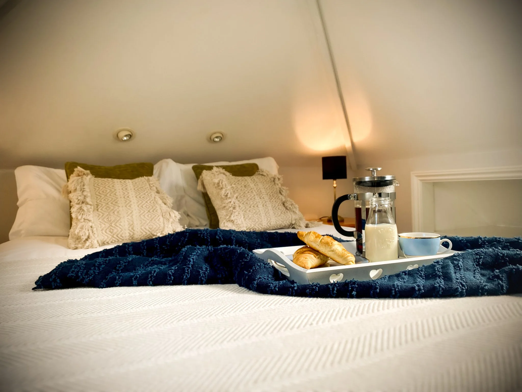 Breakfast tray with croissants, coffee, and milk on a bed with beige pillows and dark blue blanket in a cozy, lit bedroom.