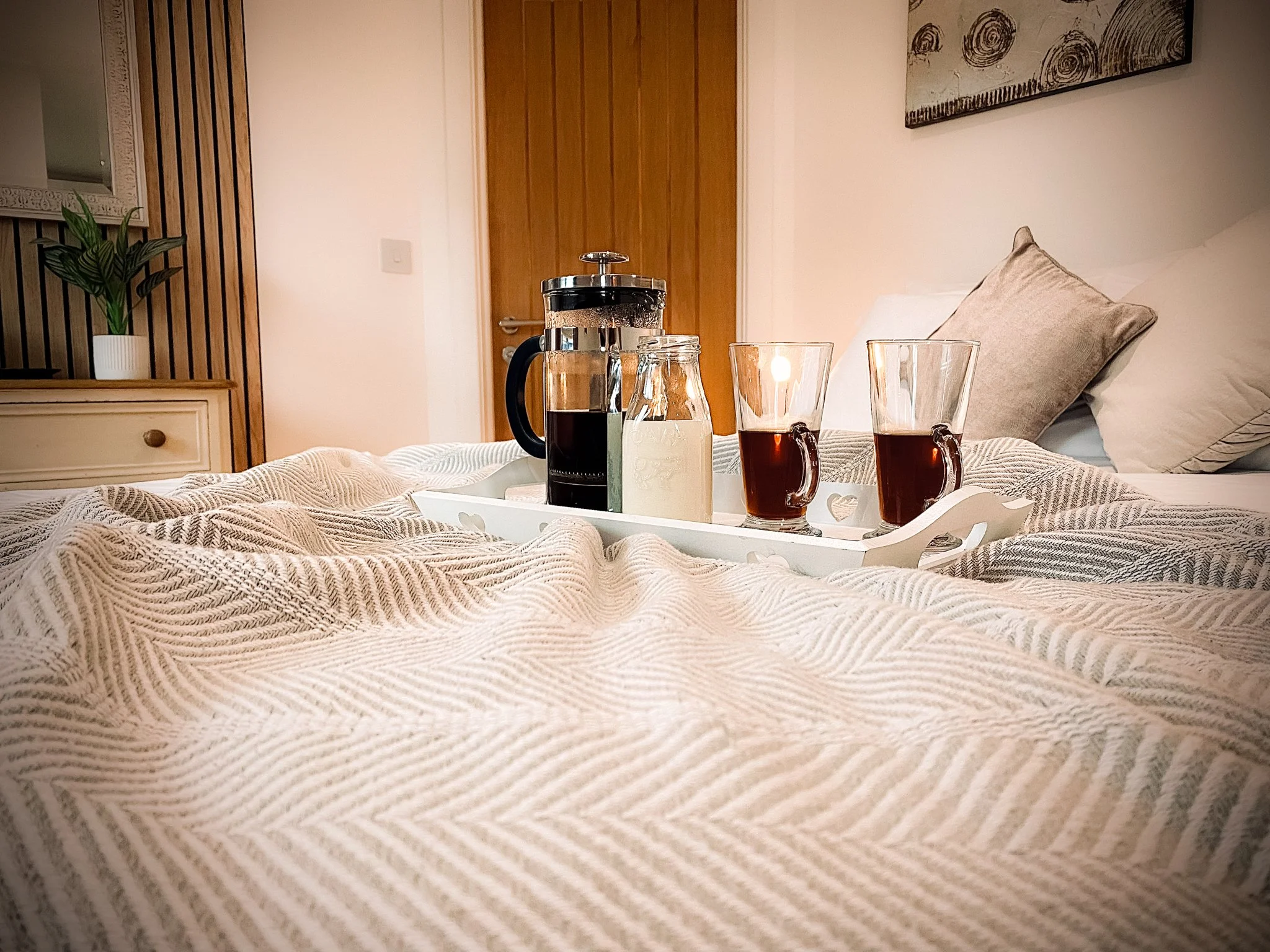 Tray with French press, milk, and two cups of coffee or tea on a bed with a textured beige blanket, pillows, and a wooden headboard. There is a potted plant and a piece of artwork on the wall.