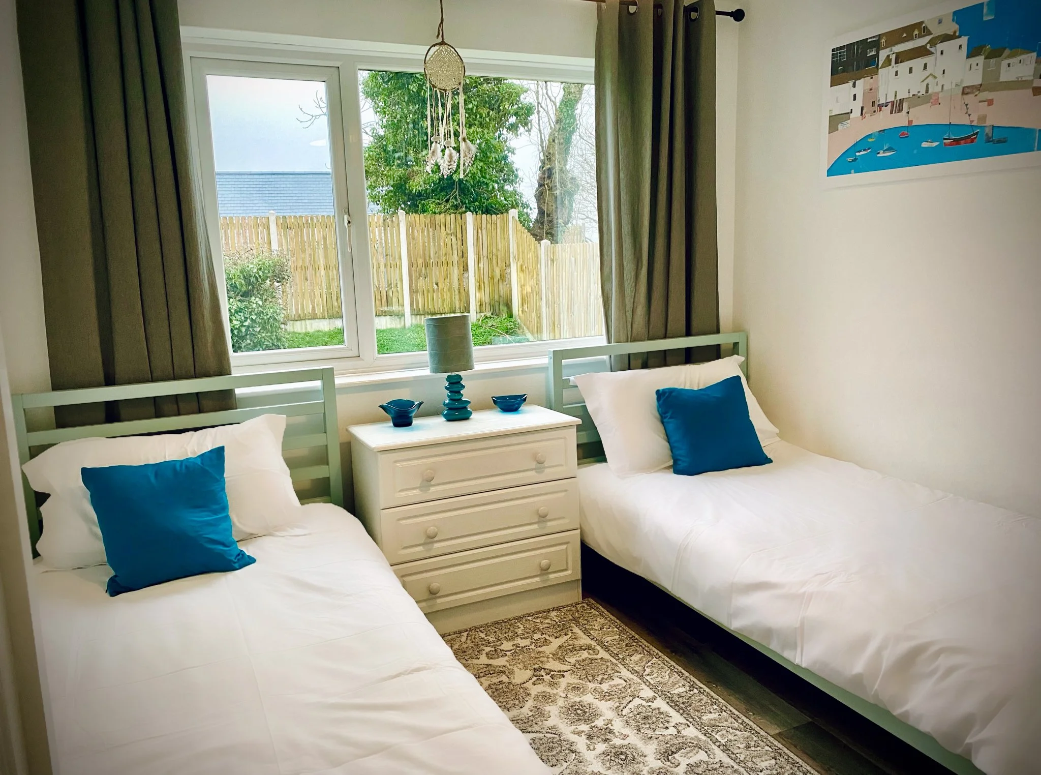 A cozy bedroom with two twin beds with white bedding and blue pillows, a white nightstand with blue decorative items and a lamp, large window with gray curtains, a patterned rug, and a colorful painting on the wall.