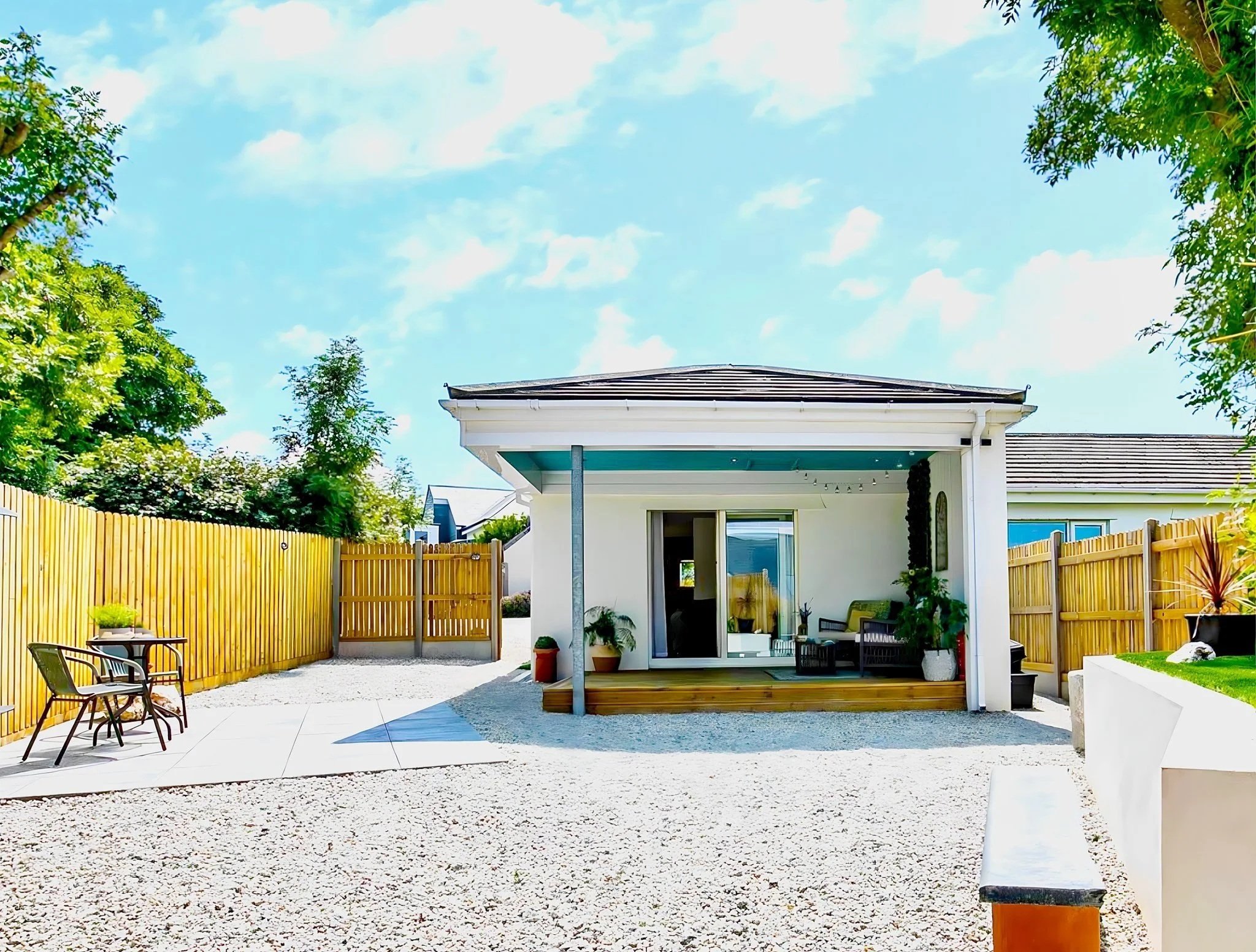 A modern backyard patio with a white house, wooden deck, and outdoor furniture, surrounded by a wooden fence and trees, under a blue sky with scattered clouds.