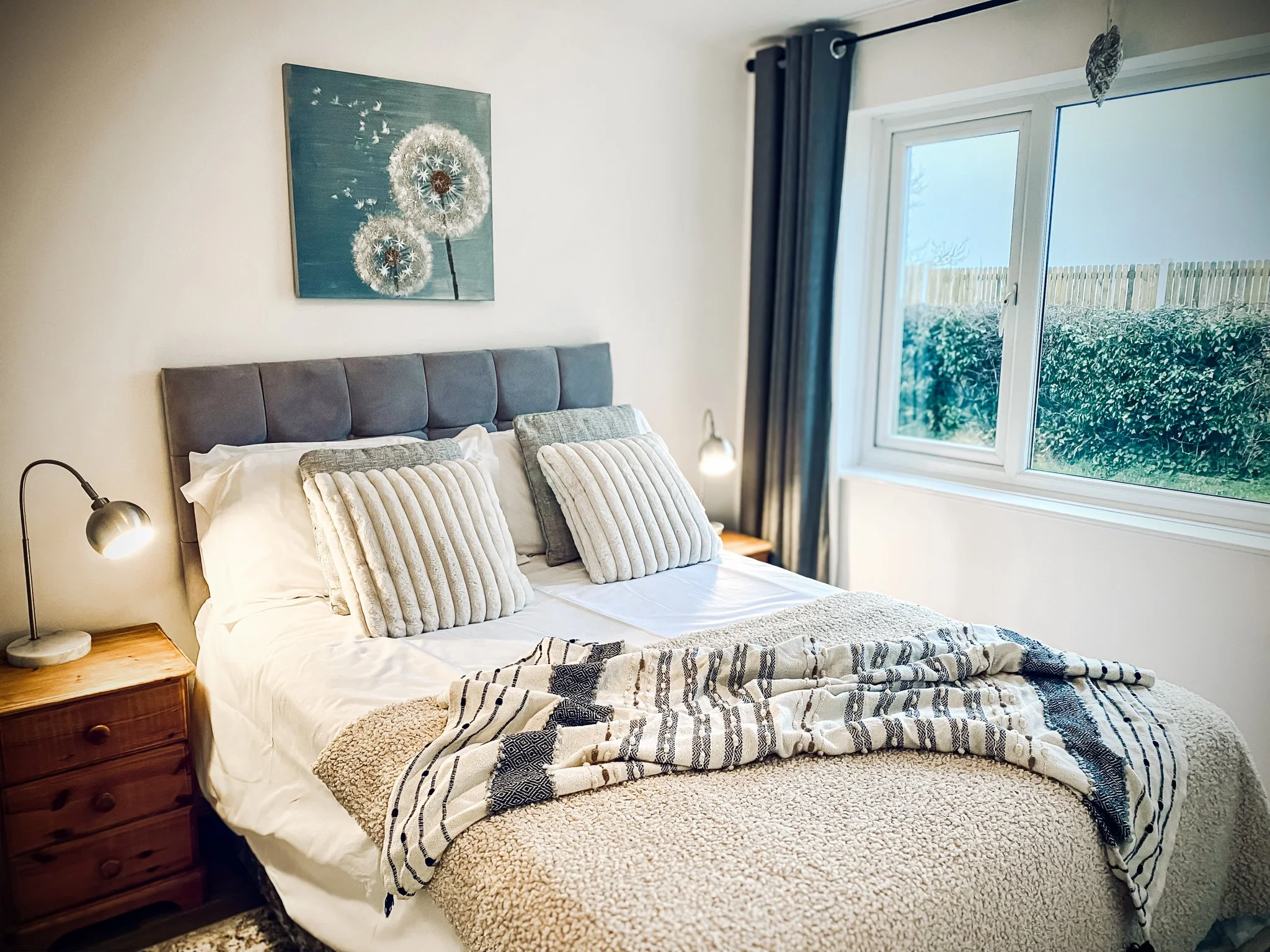 A bedroom with a bed featuring a grey upholstered headboard, six pillows, and a beige textured blanket. Two wooden nightstands with lamps flank the bed. There is a window with dark curtains, and outside greenery is visible. A dandelion floral paintin