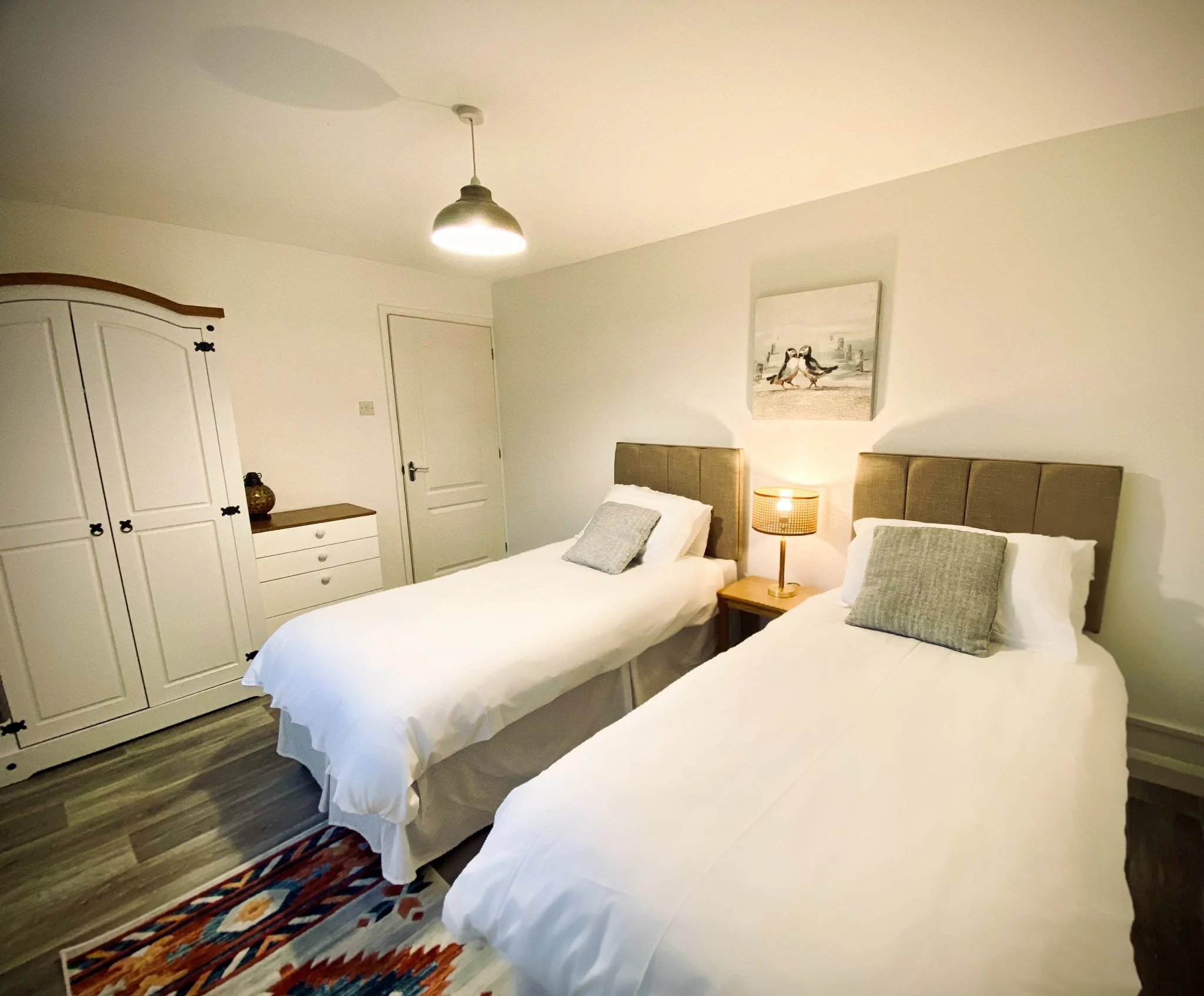 A bedroom with two single beds with beige headboards, white bedding, and gray pillows. A wooden nightstand with a lamp is between the beds. There is a white wardrobe with black handles on the left, a white dresser, and a colorful rug on the wooden fl