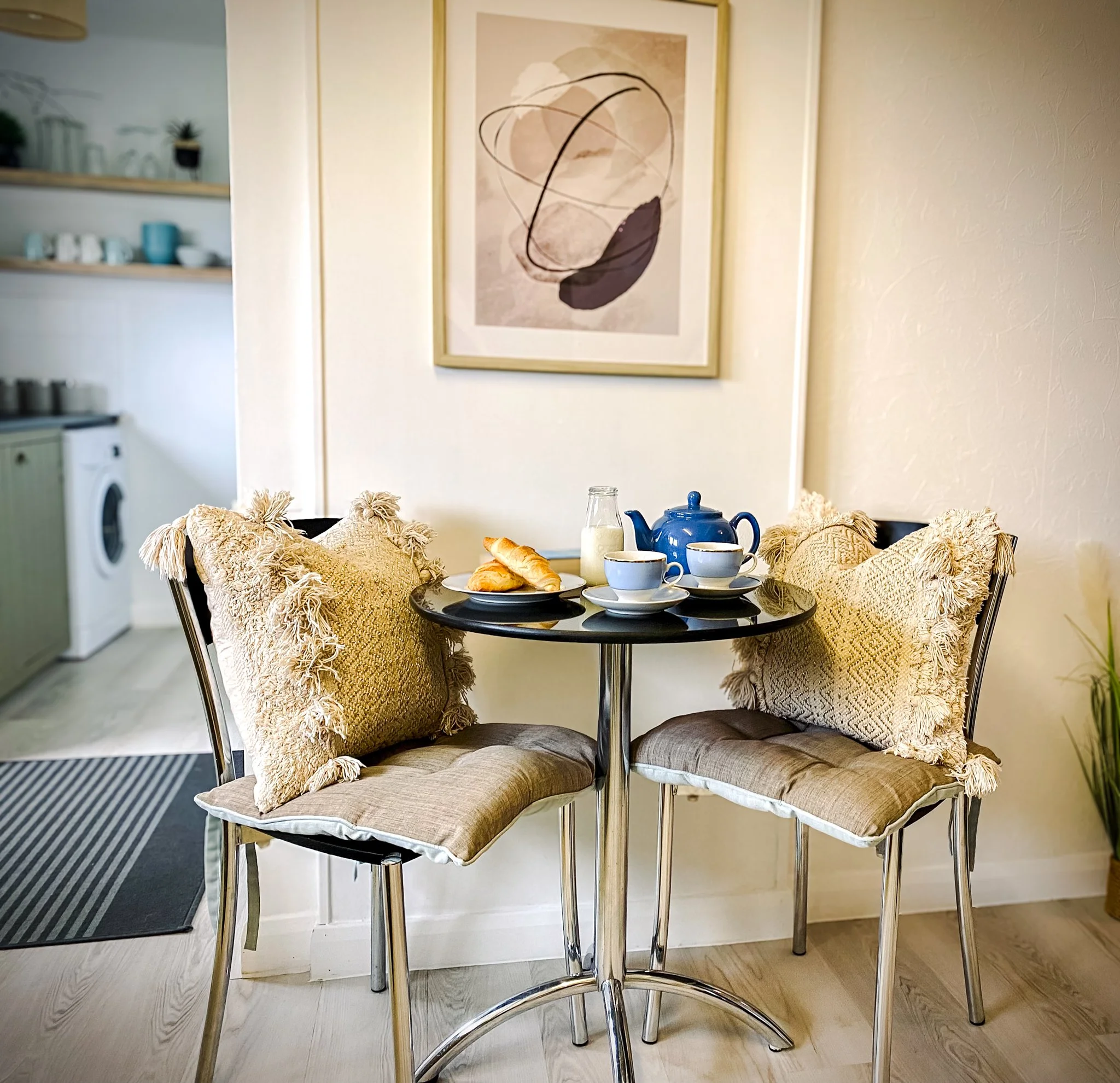 A small round dining table set for breakfast with two teacups, a teapot, a glass of milk, and croissants. There are two chairs with beige cushions and decorative pillows around the table. In the background, there is a wall with abstract artwork, a pa