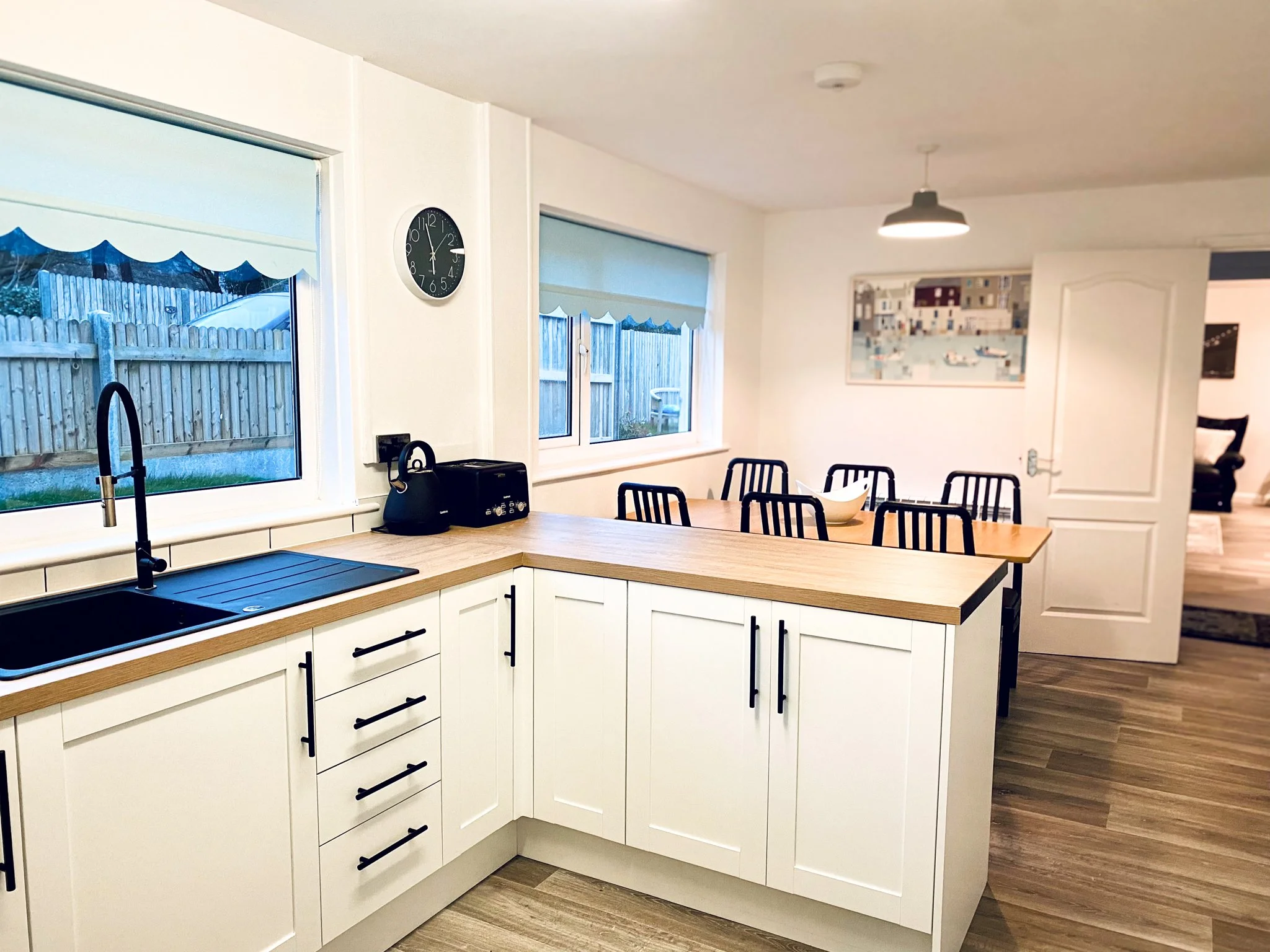 Modern kitchen with white cabinets, wooden countertops, black fixtures, and a dining area with black chairs. Two large windows with blue blinds show a backyard with a wooden fence. There is a wall clock, and a painting on the wall near the open door 