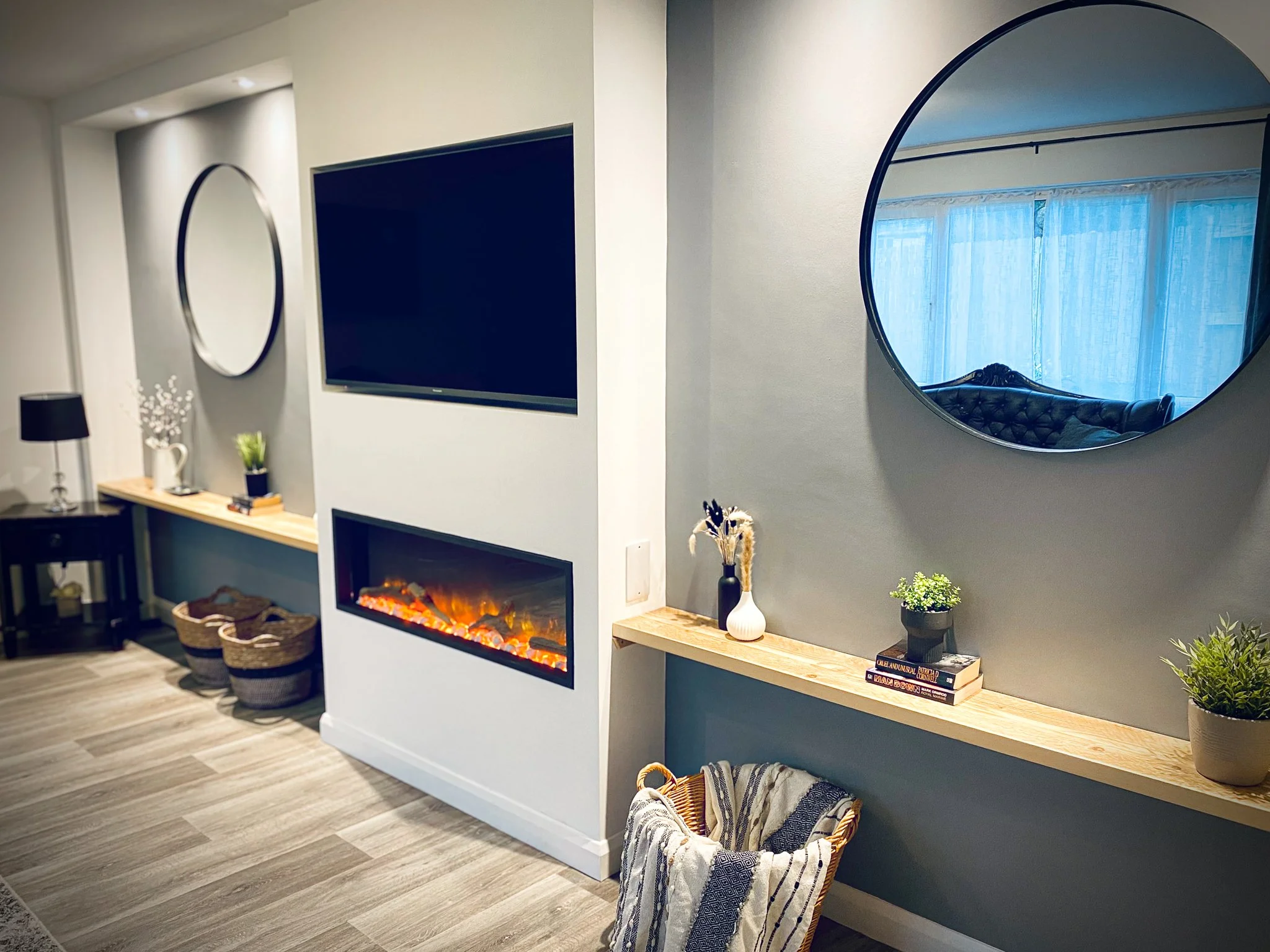 Living room with a wall-mounted flat-screen TV, an electric fireplace below it, two round mirrors on the wall, a blue painted wall, and a wooden shelf with decorative items and plants.