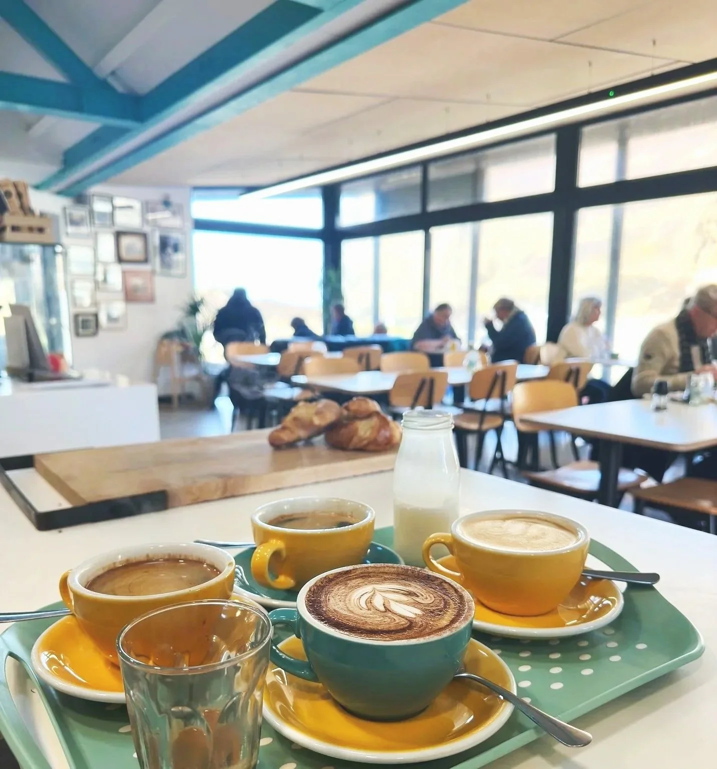 A tray with four coffee cups with different coffee drinks and a glass of water on a table in a bright cafe with large windows, a milk bottle, and some pastries in the background.