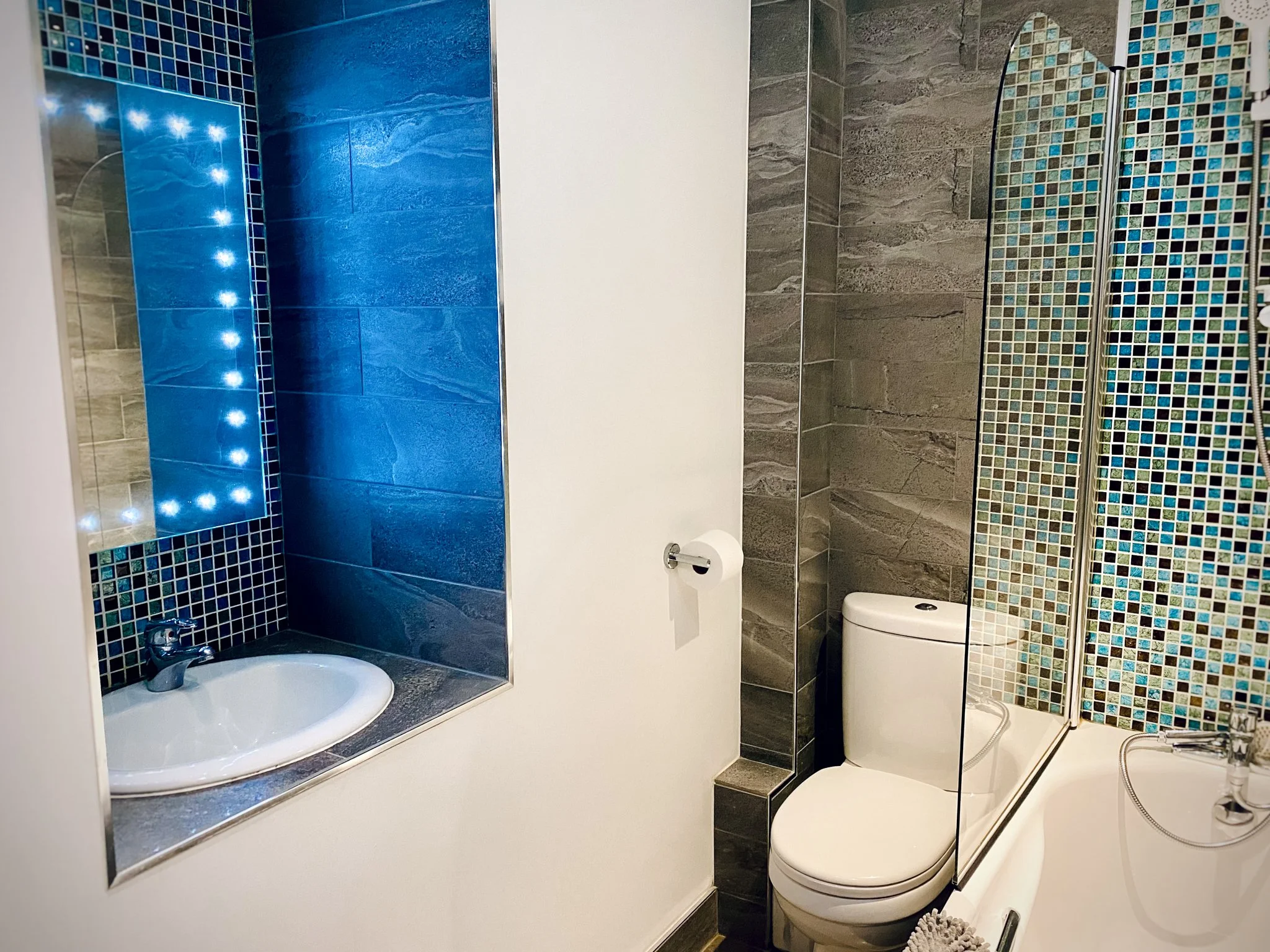 Bathroom with a mirror with LED lights, blue and gray tile walls, a small sink, a toilet, and a bathtub with a handheld showerhead.