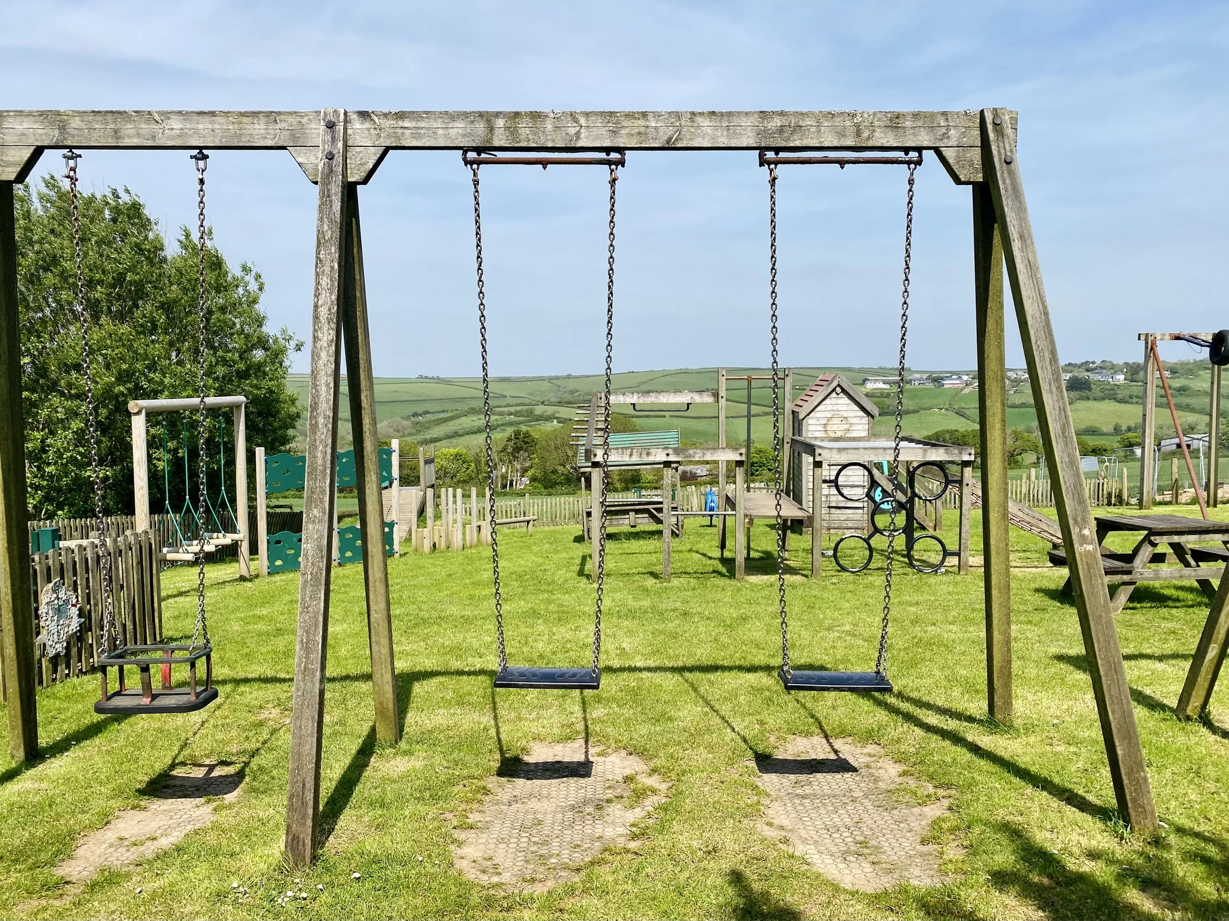 Empty outdoor playground with swings, climbing structures, and picnic tables on a grassy field under a cloudy blue sky, with hills in the background.